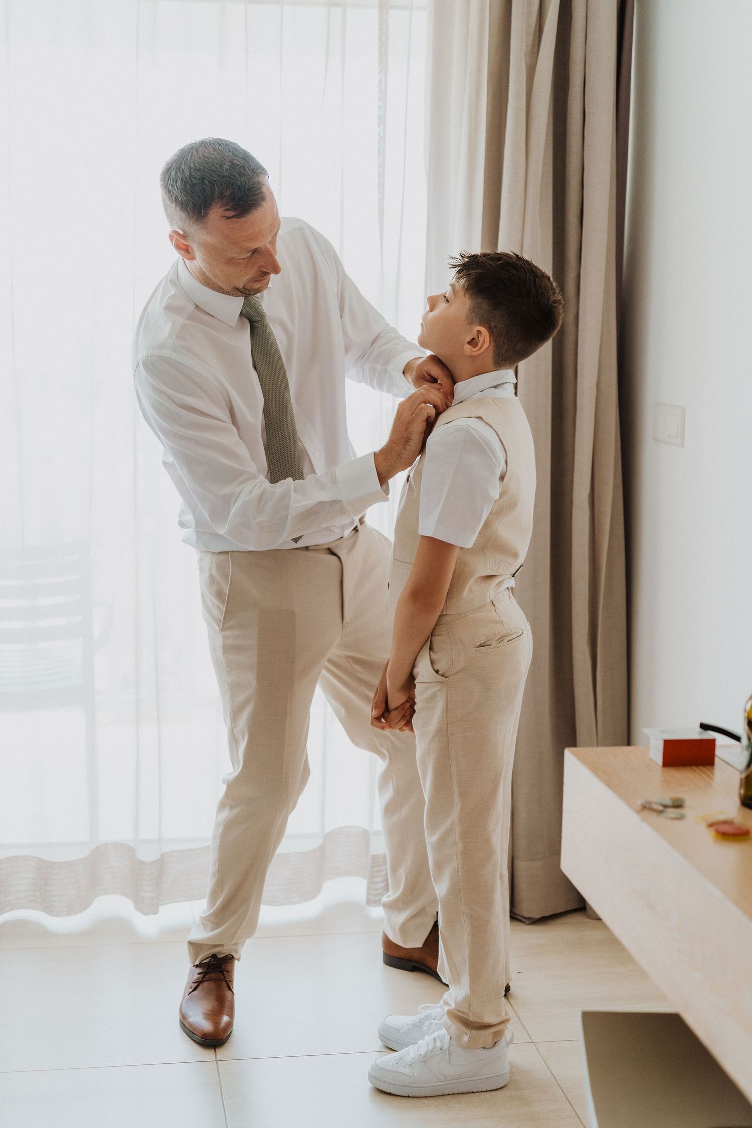A father in white dress shirt helps his young son adjust his tie while getting ready in a bright modern room.
