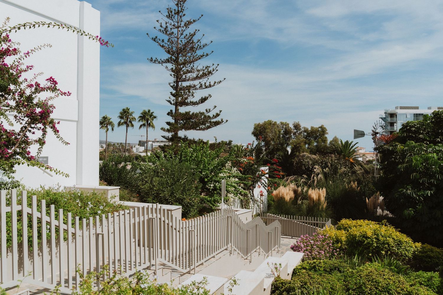 White gated terrace with palm trees and tropical landscaping against a sunny blue sky in a coastal setting.