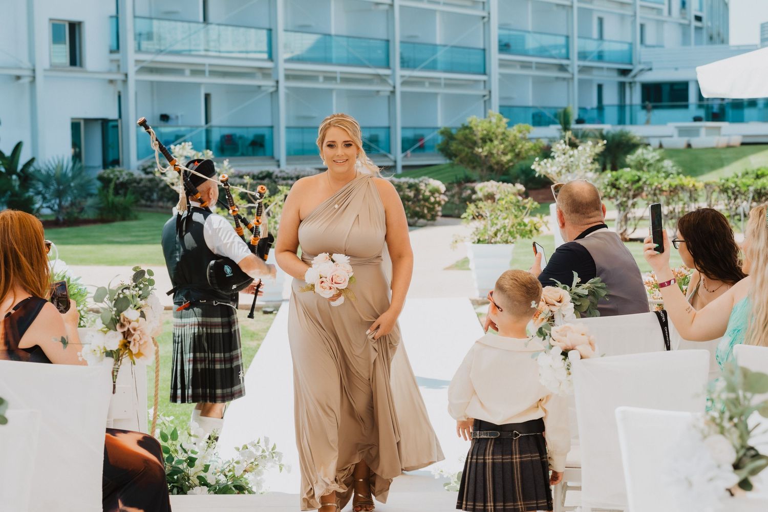 Wedding processional with bagpiper and attendants walking down aisle with white chairs.