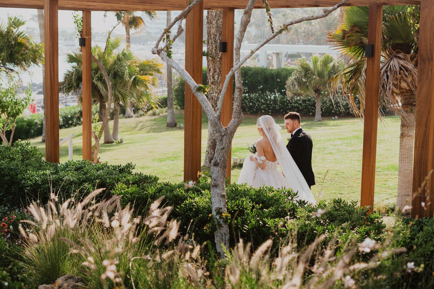 Romantic wedding portrait through wooden pergola frames with lush garden landscaping.