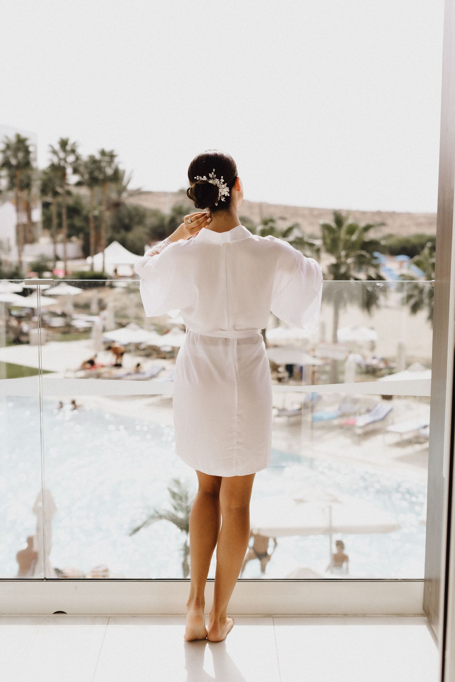 Person in white robe standing on balcony overlooking resort pool and palm trees.