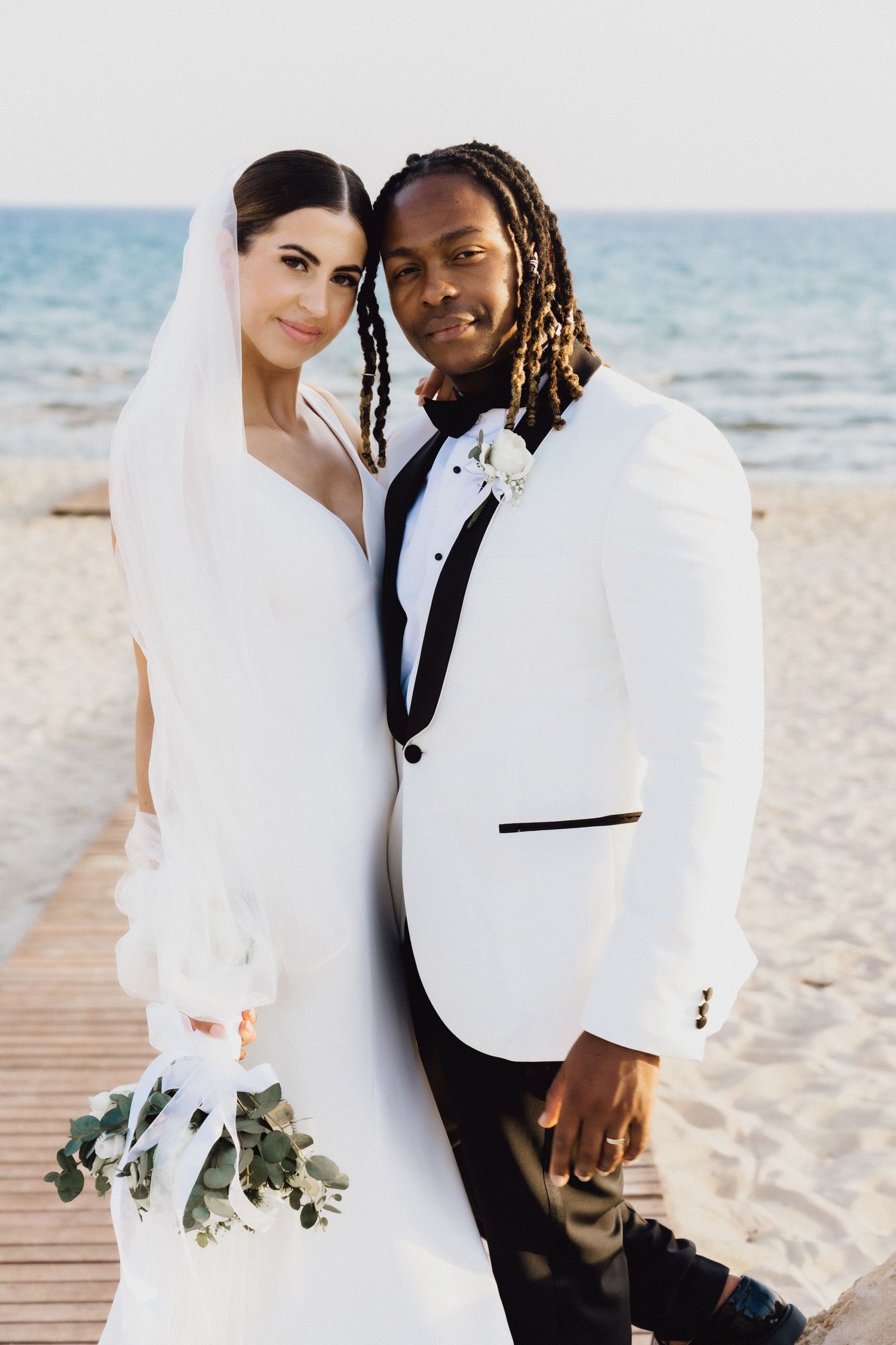 A bride and groom pose together on a beach boardwalk, with the groom wearing a white tuxedo jacket with black trim.