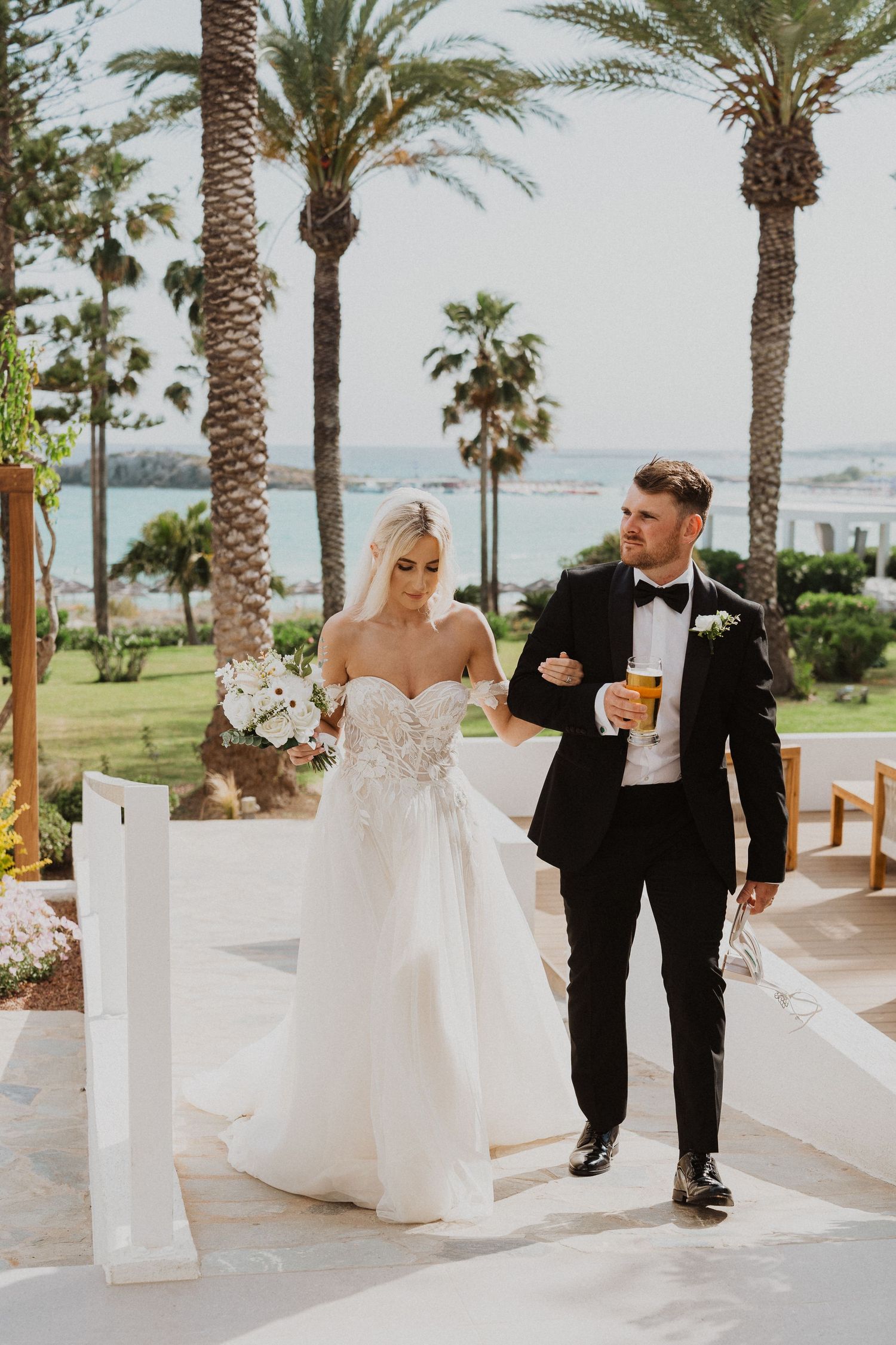 Newlyweds stroll along a palm-lined pathway at an oceanfront wedding venue.
