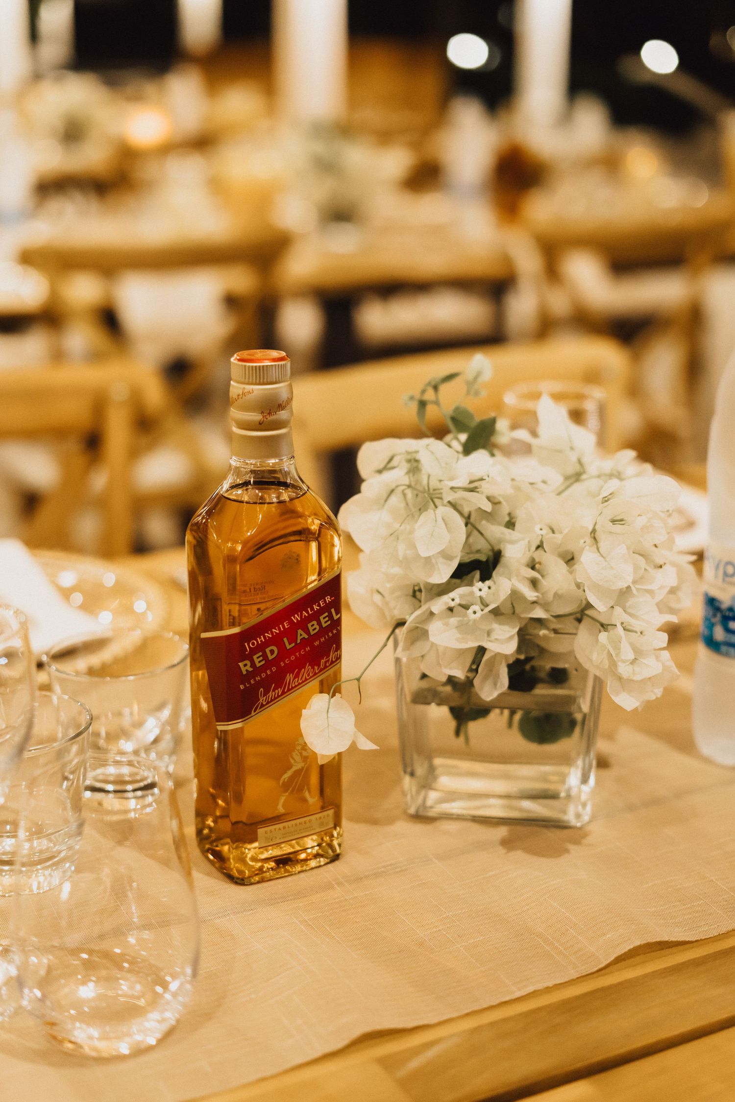 A table setting with a Johnnie Walker bottle and white floral centerpiece at a wedding reception.