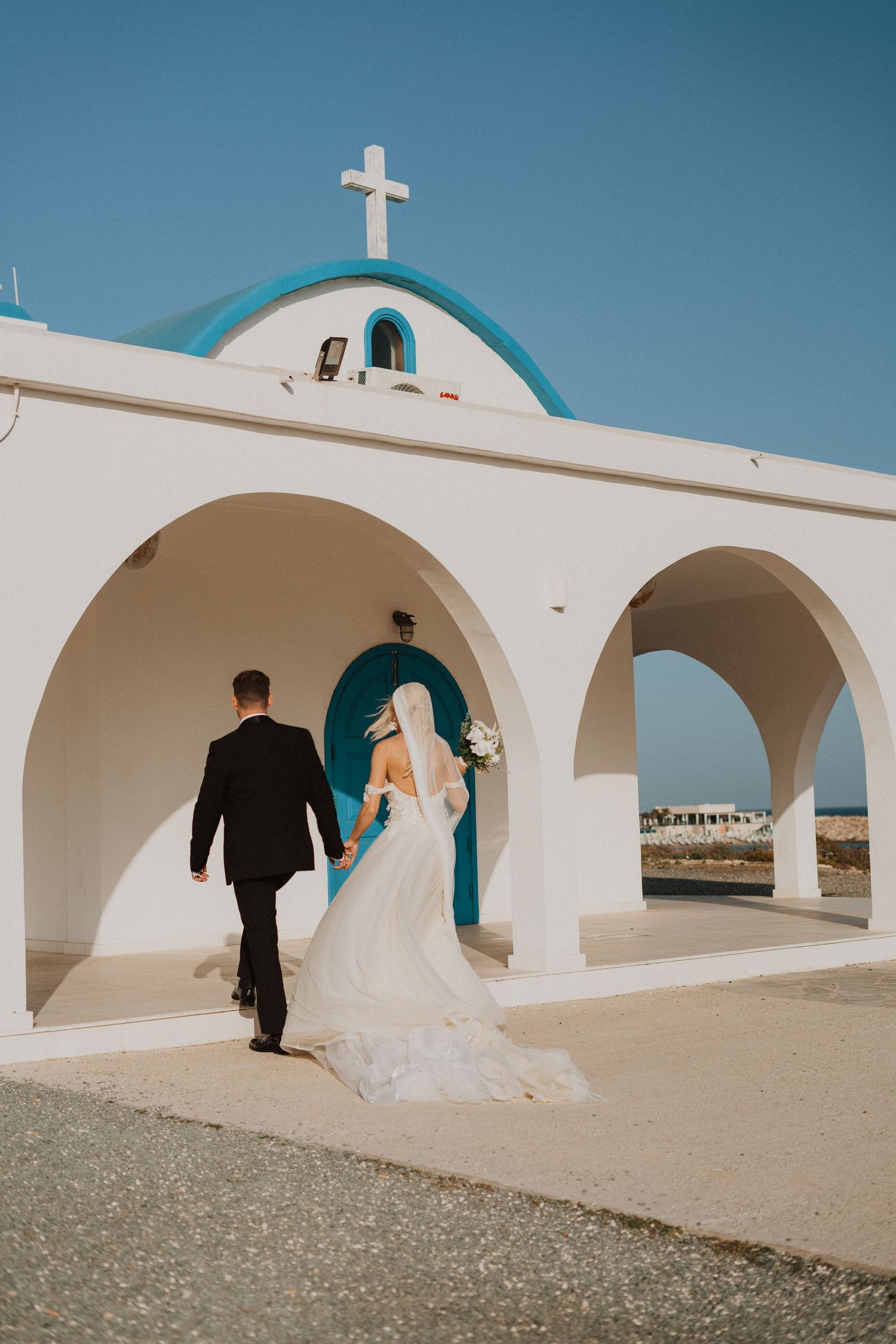 Wedding couple enters white Mediterranean chapel with blue dome and cross.