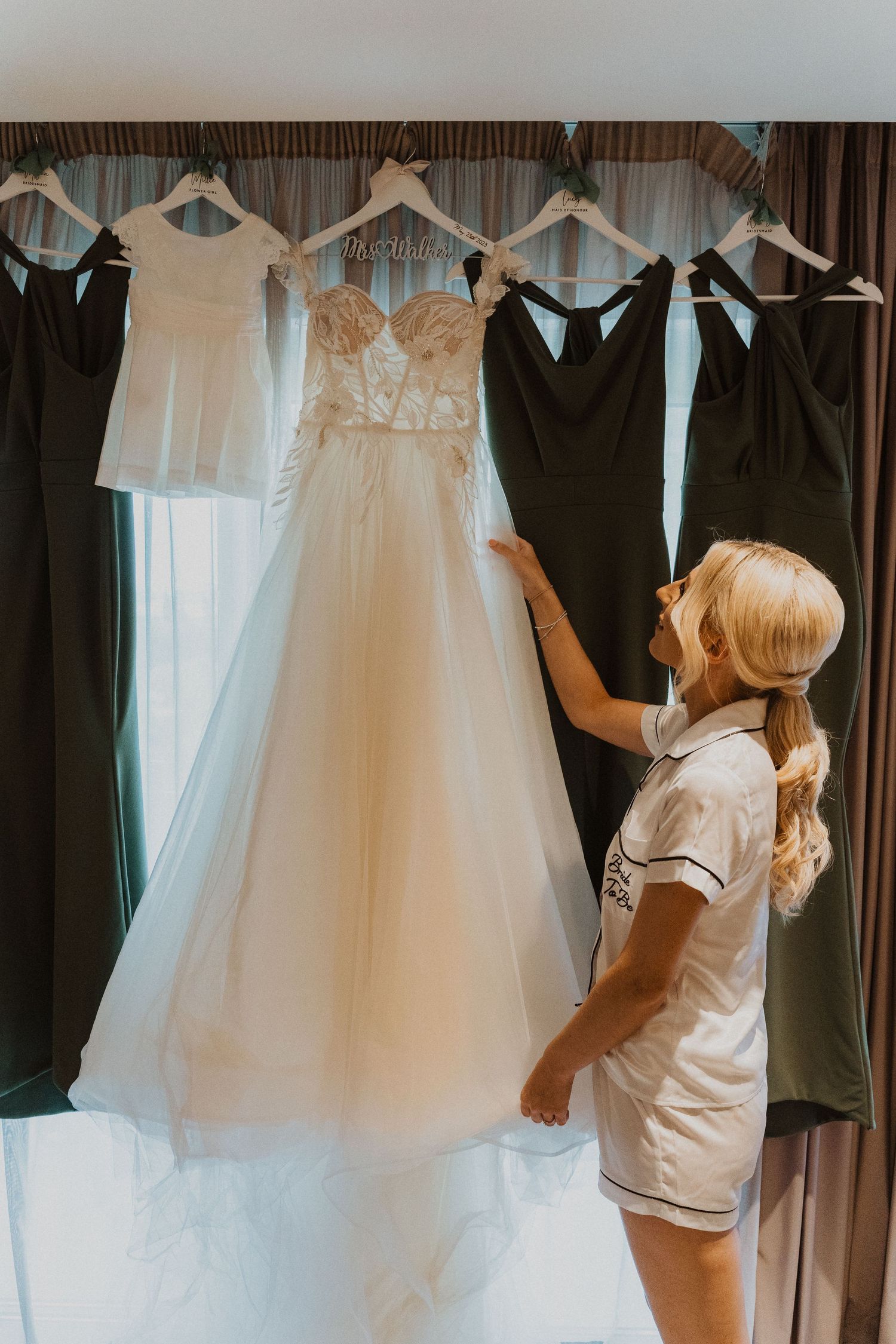 Wedding dresses and bridesmaid gowns displayed on hangers against brown curtains.