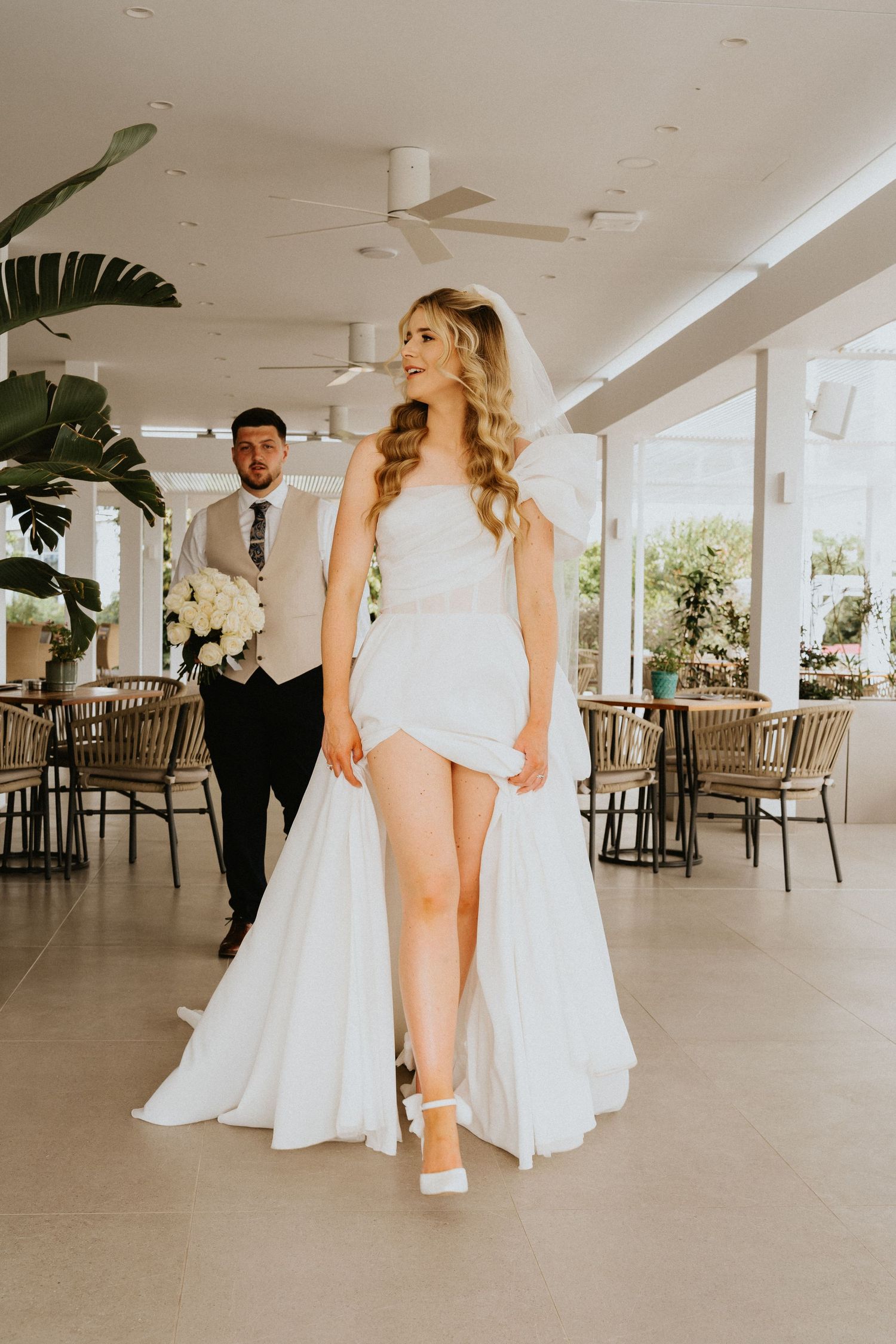 Elegant bride in split dress poses in modern indoor venue with palm decor.
