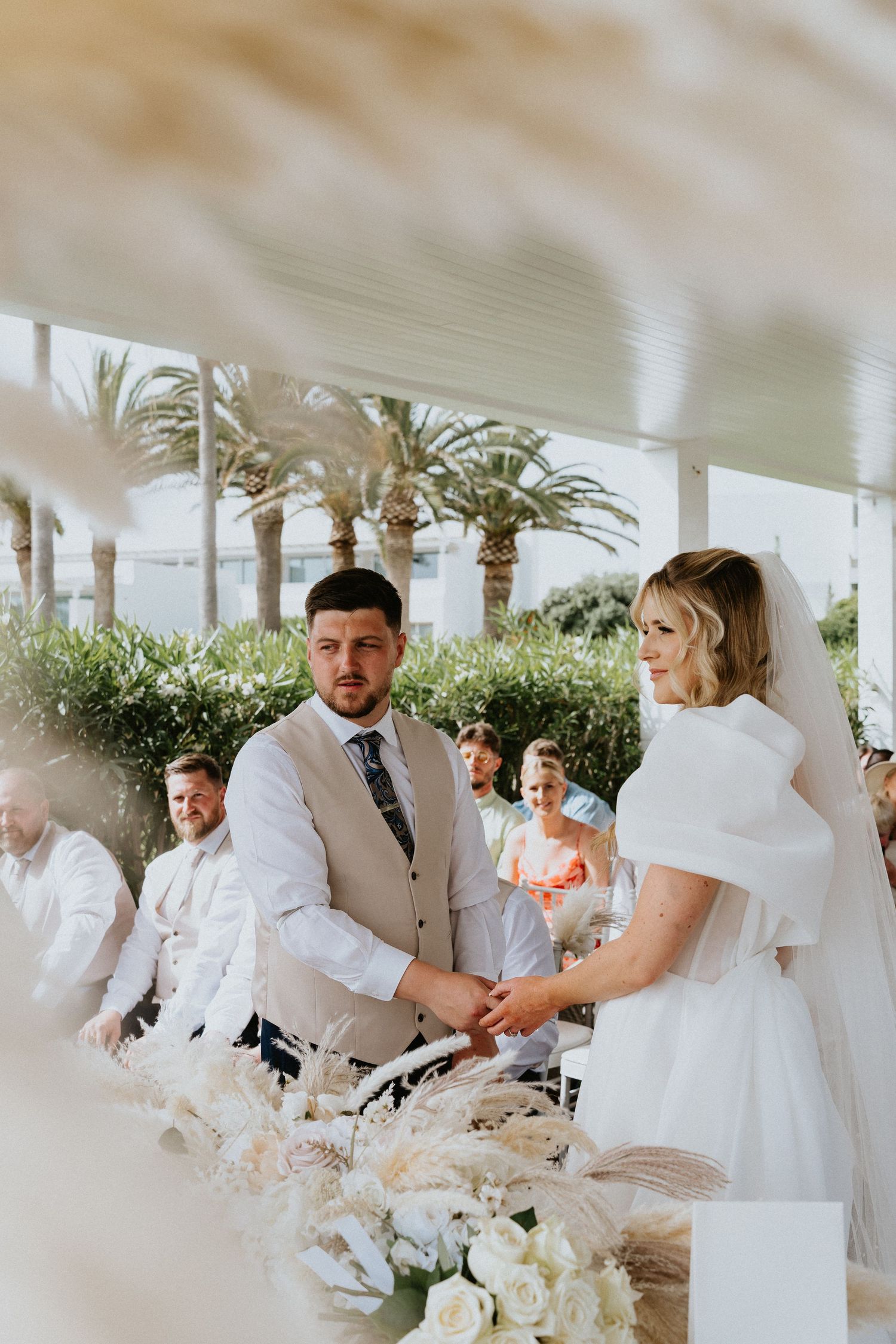 Couple exchanges vows during outdoor ceremony with white floral decorations and palm trees.