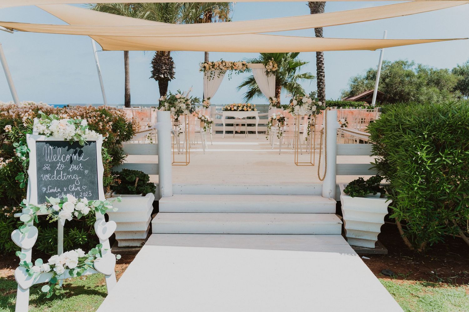 An elegant outdoor wedding ceremony setup with white stairs, floral decorations, and draped fabric canopy against a garden backdrop.