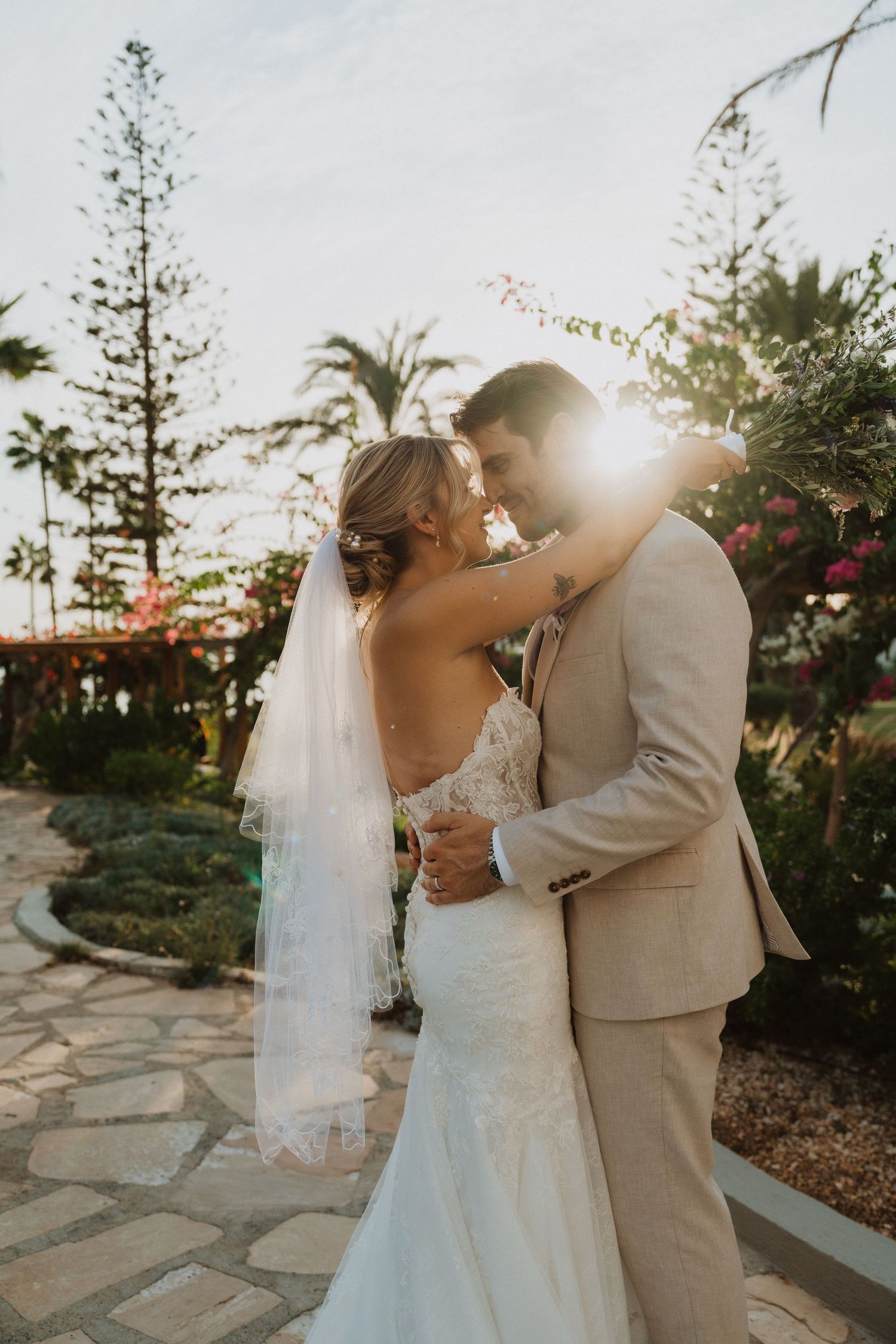 Sun flare illuminates romantic wedding portrait on stone pathway with tropical landscaping.