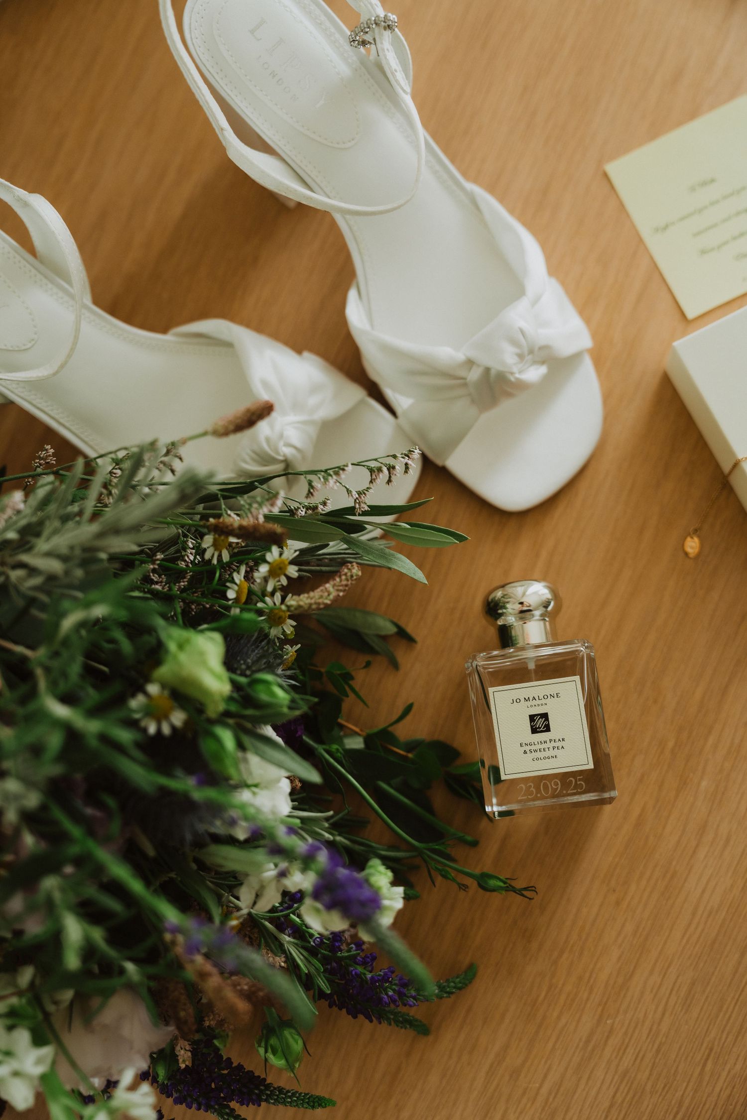 White bridal shoes and Jo Malone perfume arranged with fresh flowers on wooden floor.