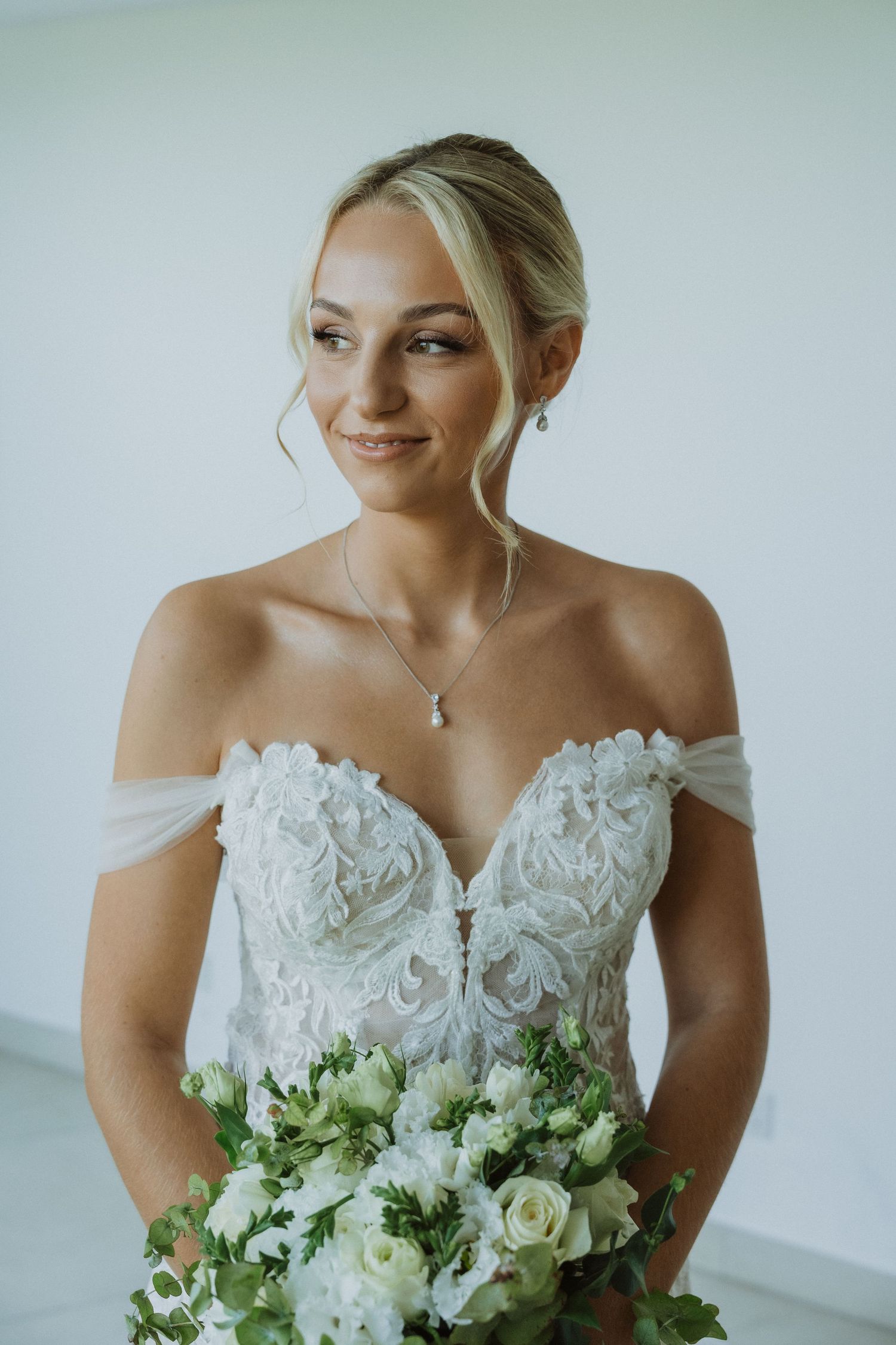 Bride smiling while holding a fresh white and green bouquet in an off-shoulder wedding dress.