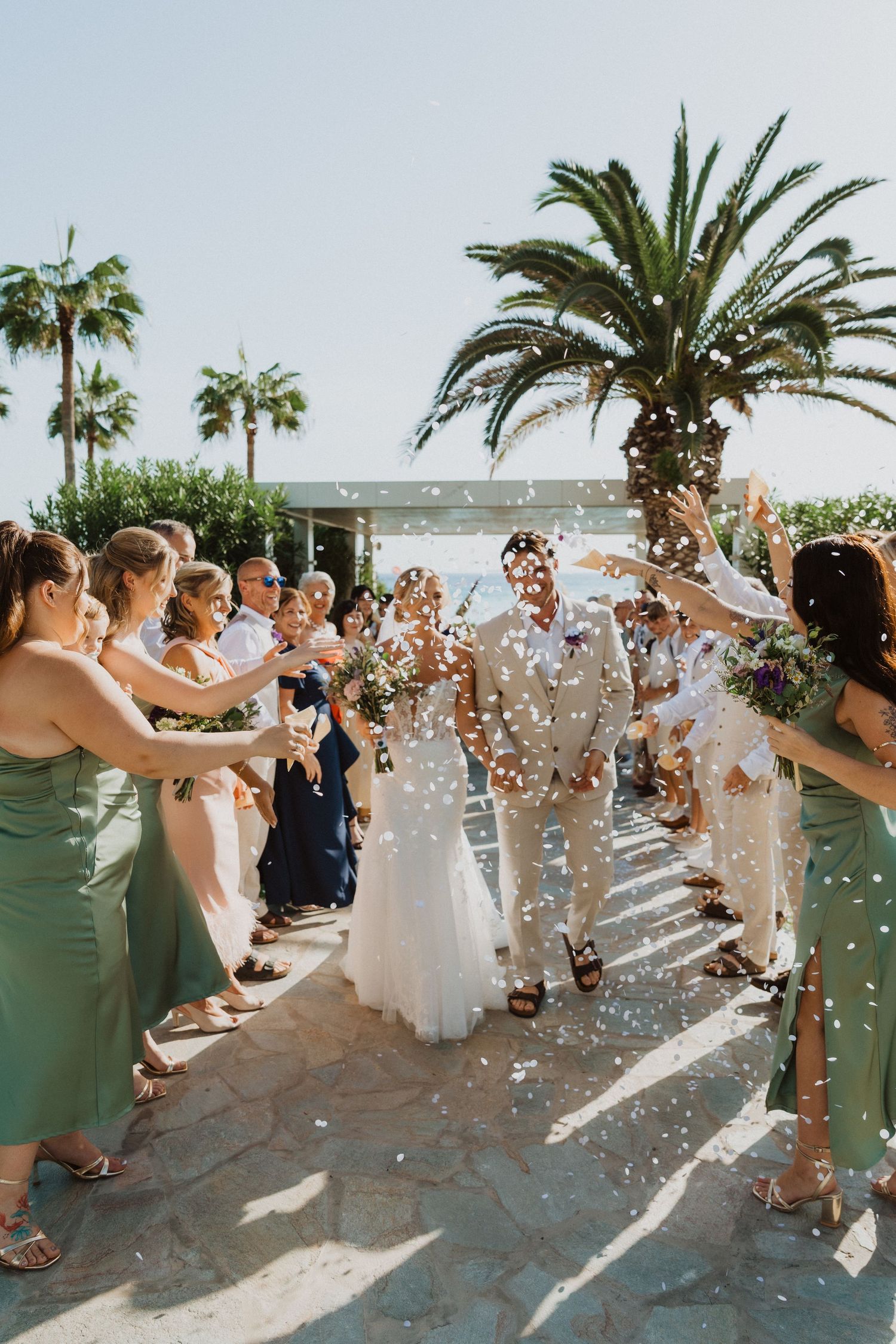 Joyful wedding exit with guests throwing confetti under tropical palm trees.