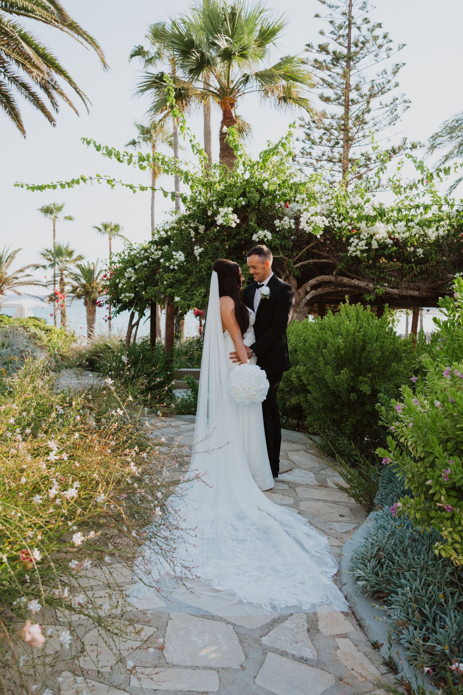 A wedding couple poses on a garden path with palm trees and flowering bushes in the background.