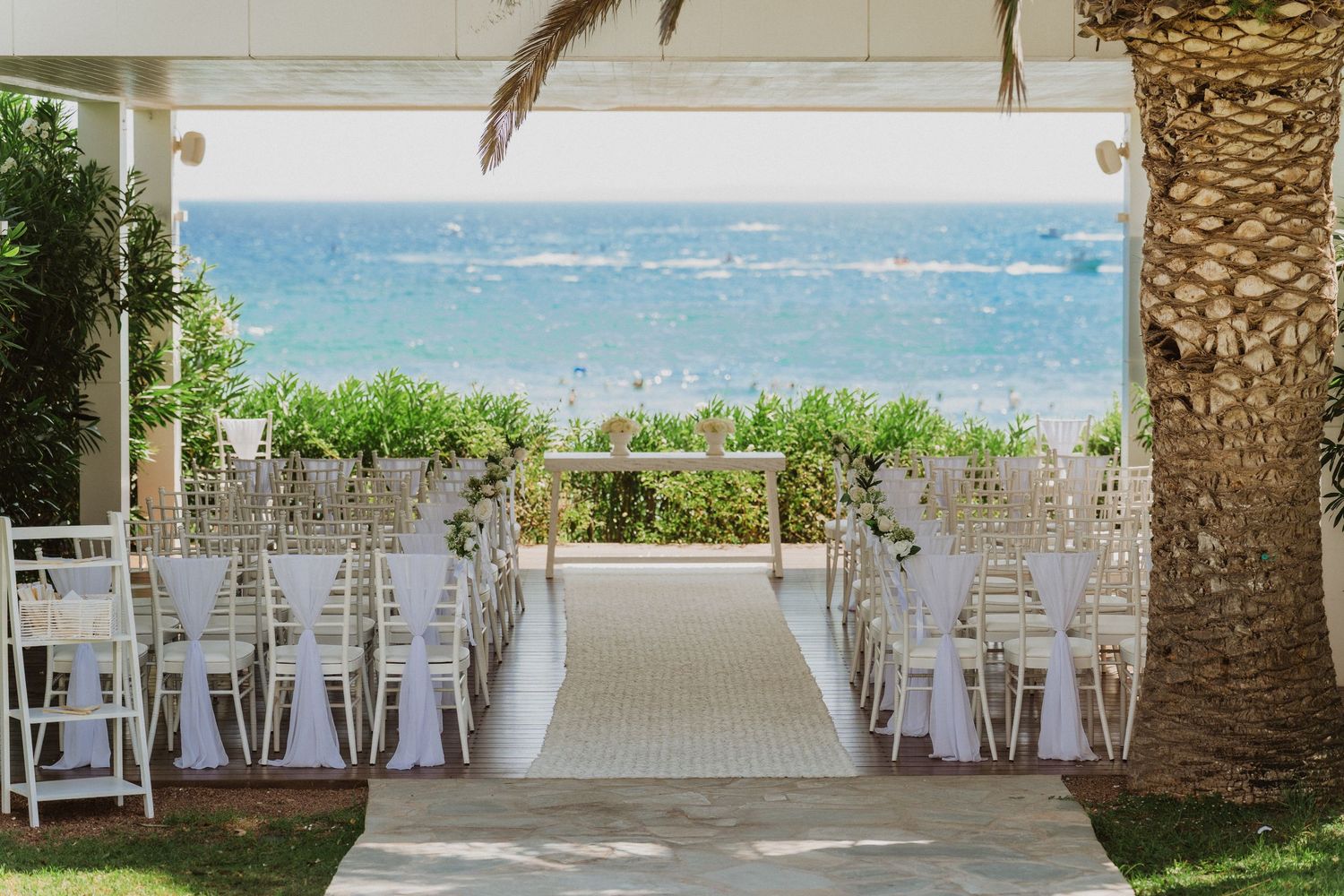 White chairs arranged for a beach wedding ceremony with palm trees and ocean views in the background.