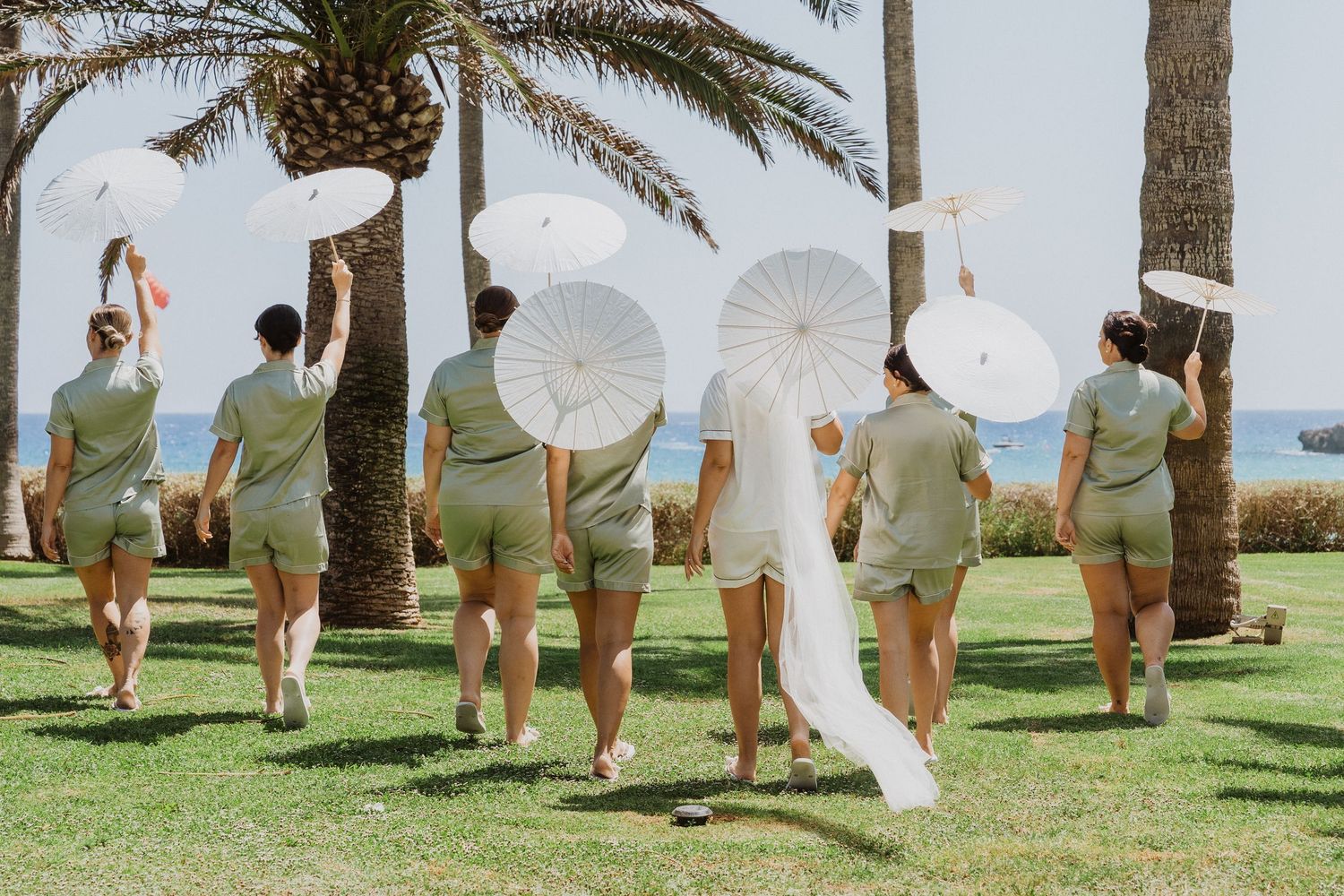 Wedding party members walk in line across a tropical beach lawn holding white parasols on a sunny day.