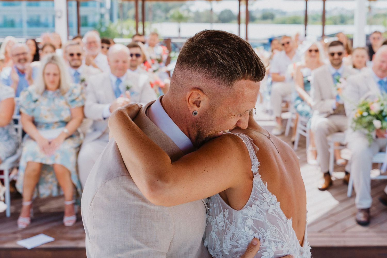 A couple shares a romantic kiss during their beachfront wedding ceremony with guests watching from white chairs.