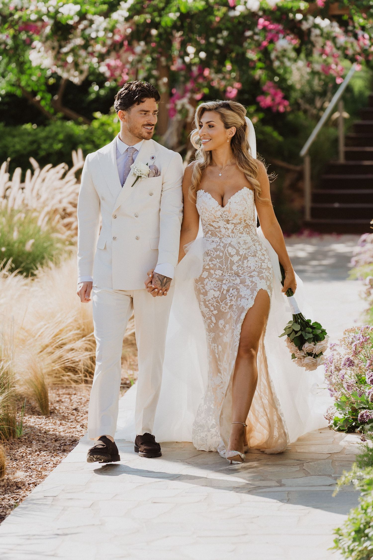 A couple walking together outdoors on their wedding day, wearing a white suit and lace wedding gown with slit.