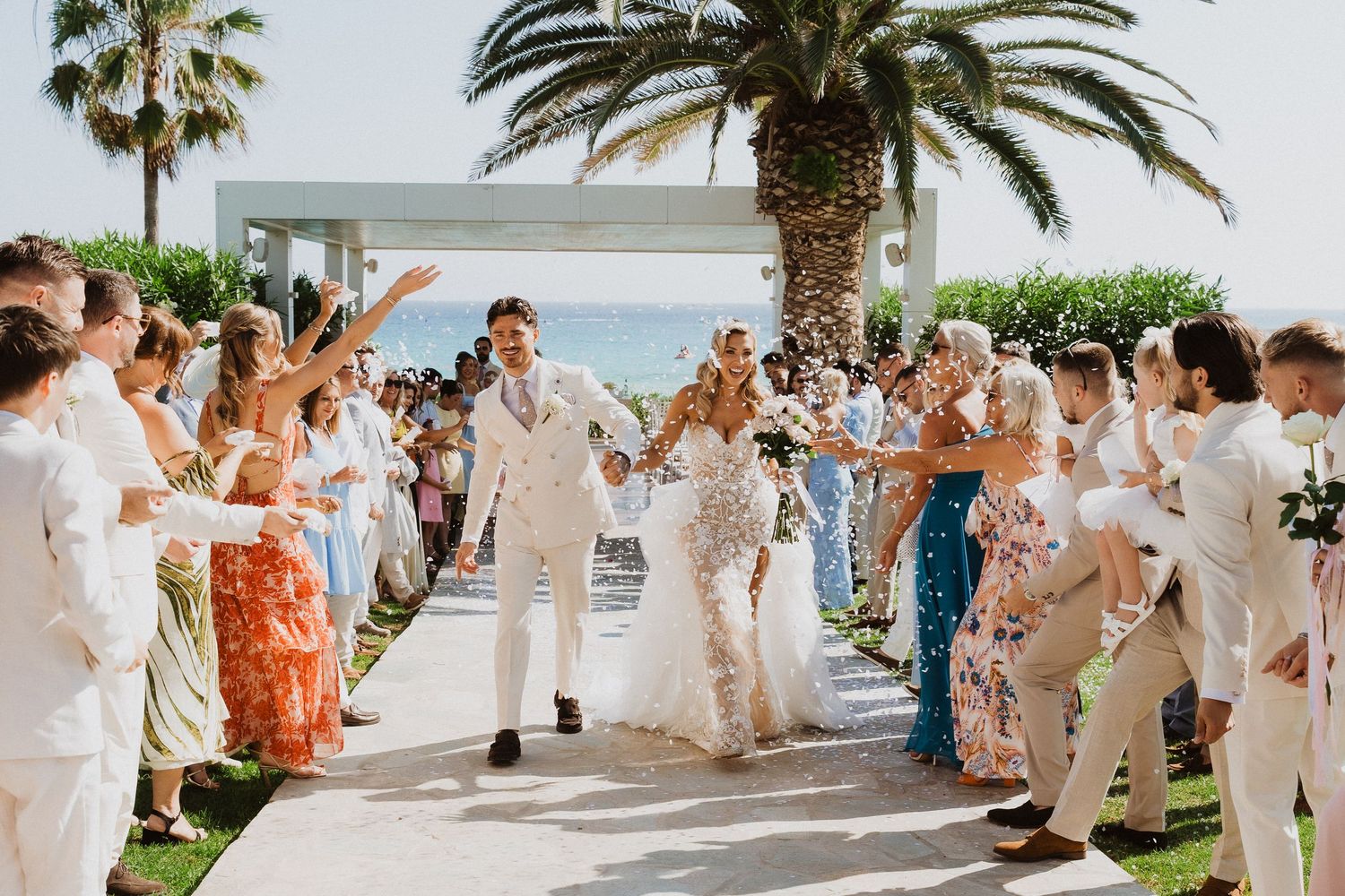 Wedding guests in white attire line a palm tree-lined walkway as newlyweds joyfully exit their beachside ceremony.
