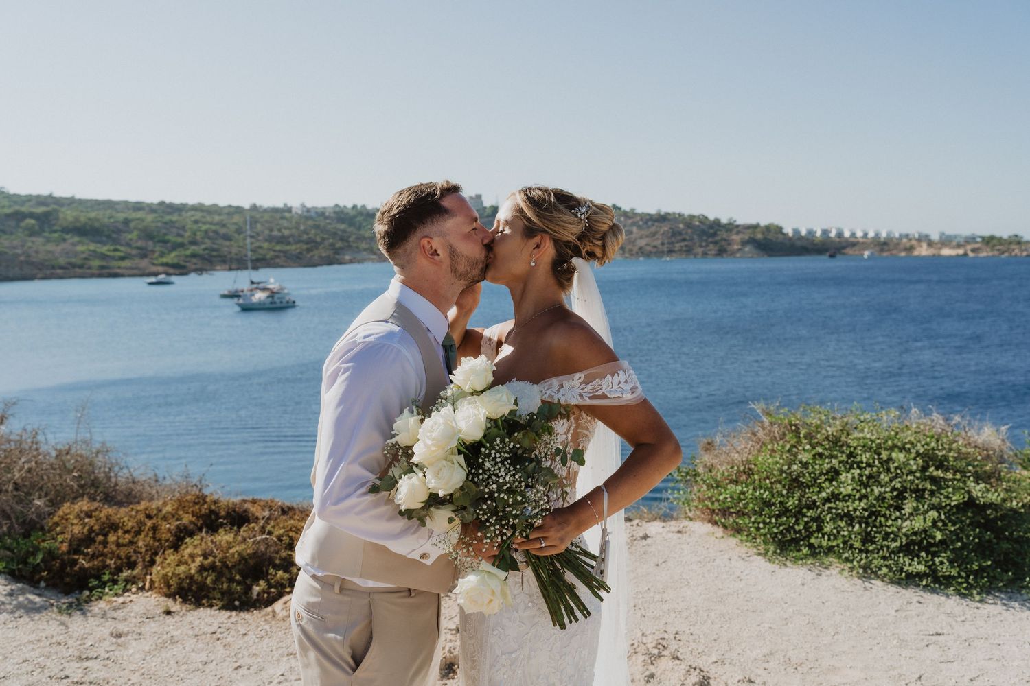 A romantic seaside wedding ceremony with a couple embracing on a sunny beach overlooking the ocean.