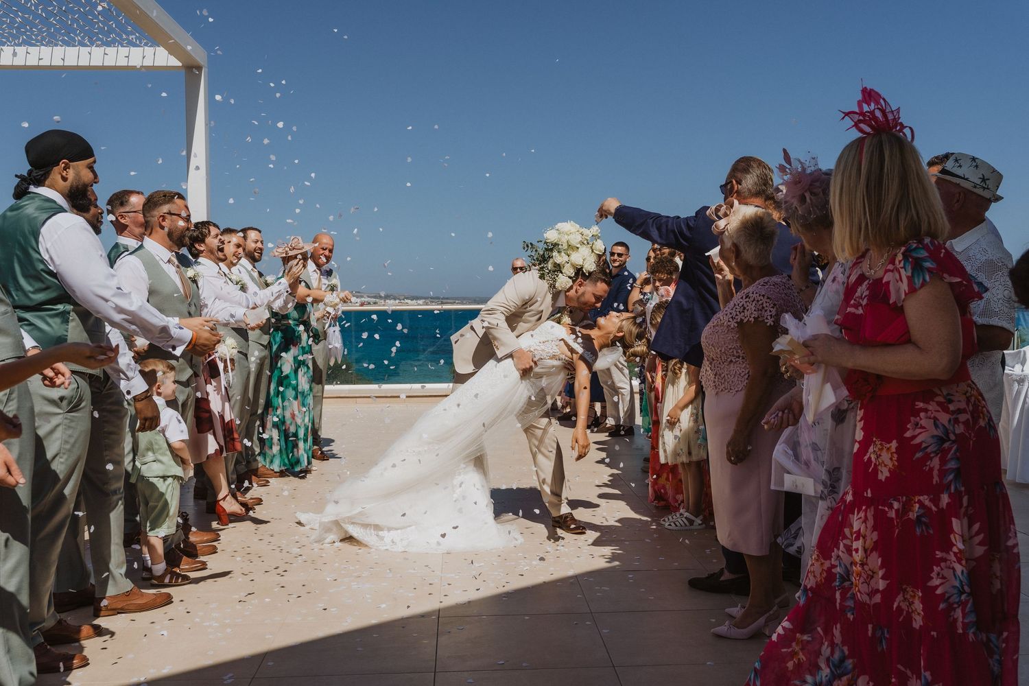 Wedding guests throw confetti on a sunny beach as a couple walks between two rows of celebrants by the ocean.