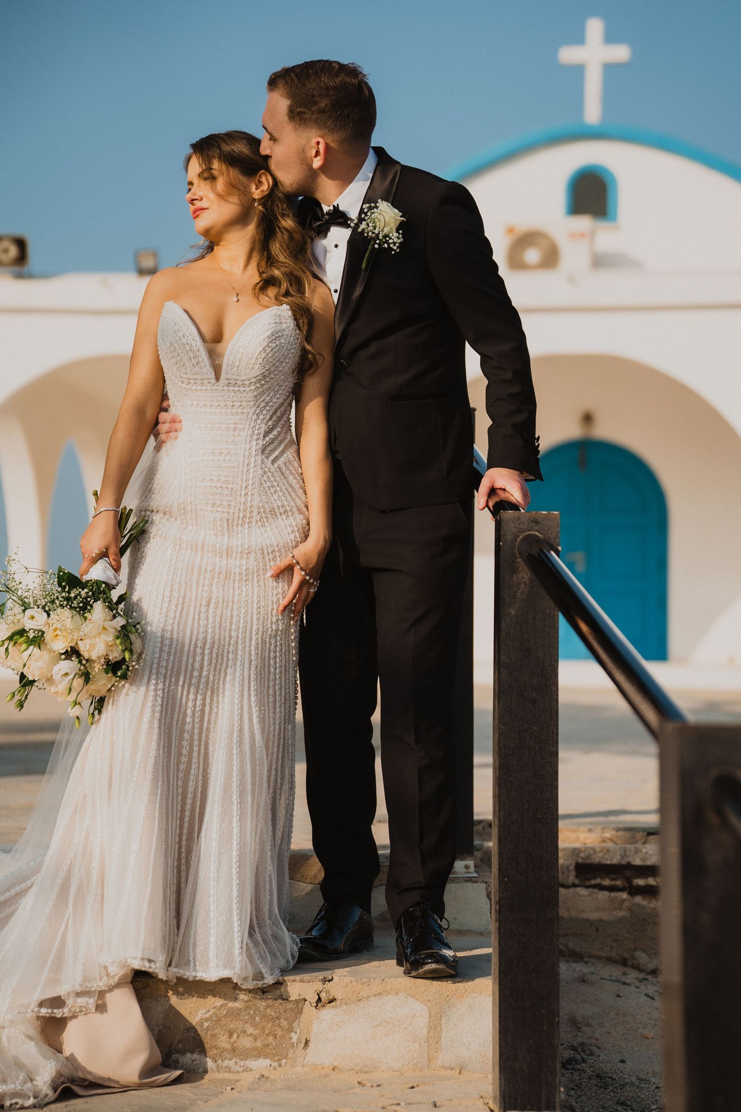Bride and groom pose together near a white chapel with blue accents under a clear sky.
