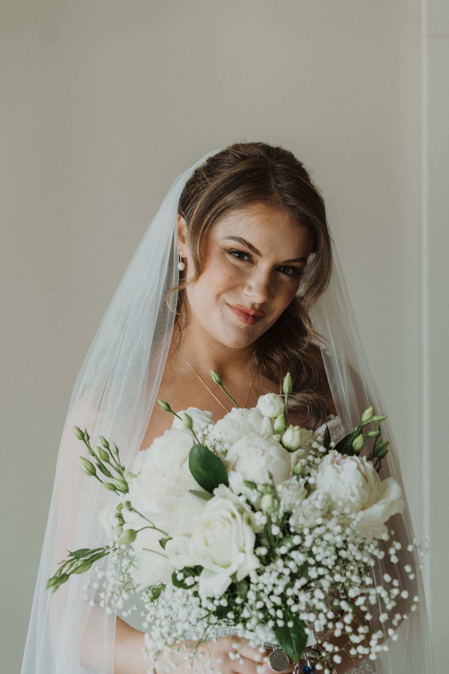 Beautiful bride wearing a veil and holding a white floral bouquet smiling softly.