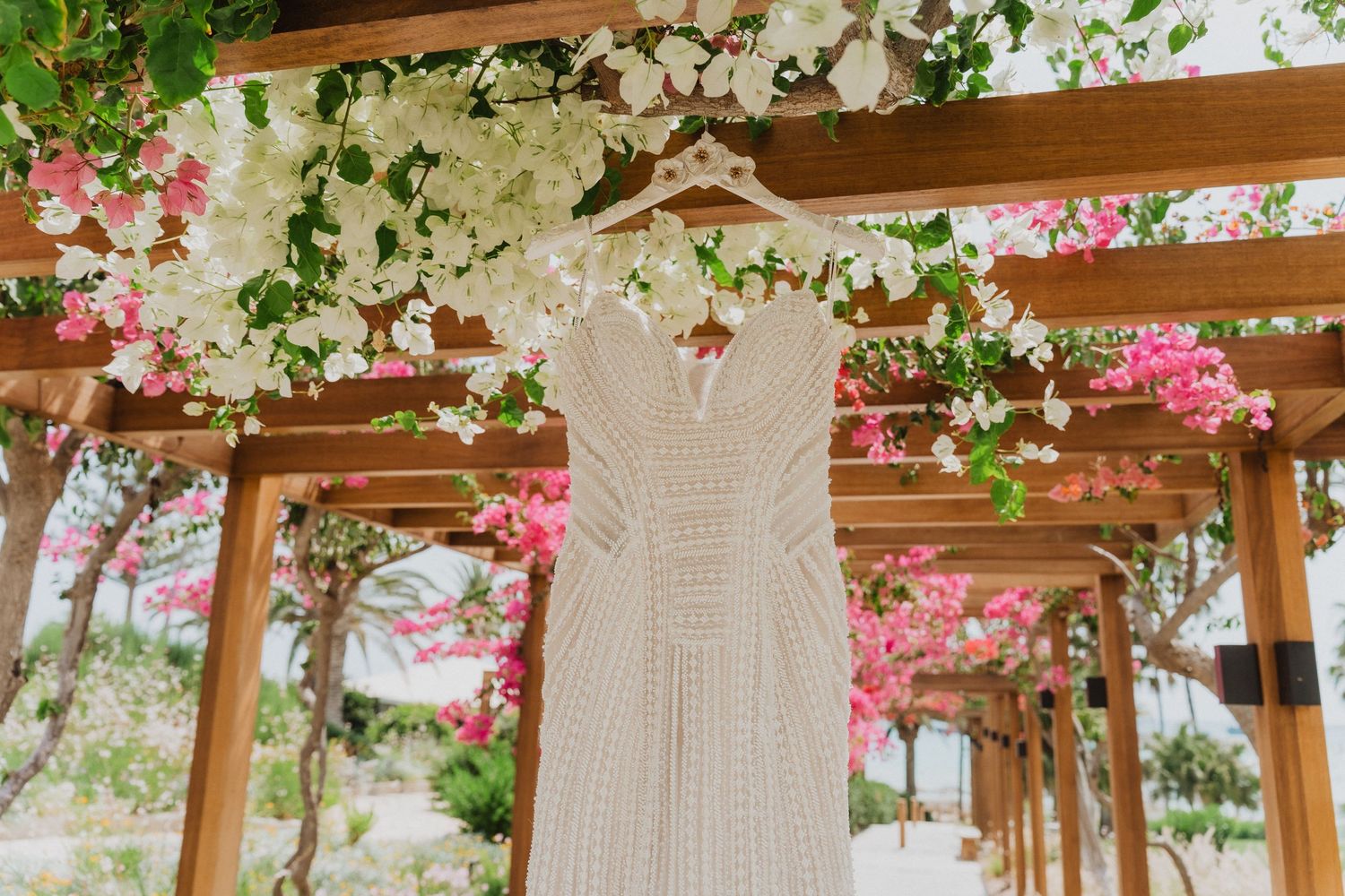 White lace wedding dress hanging on wooden pergola adorned with pink and white bougainvillea flowers.