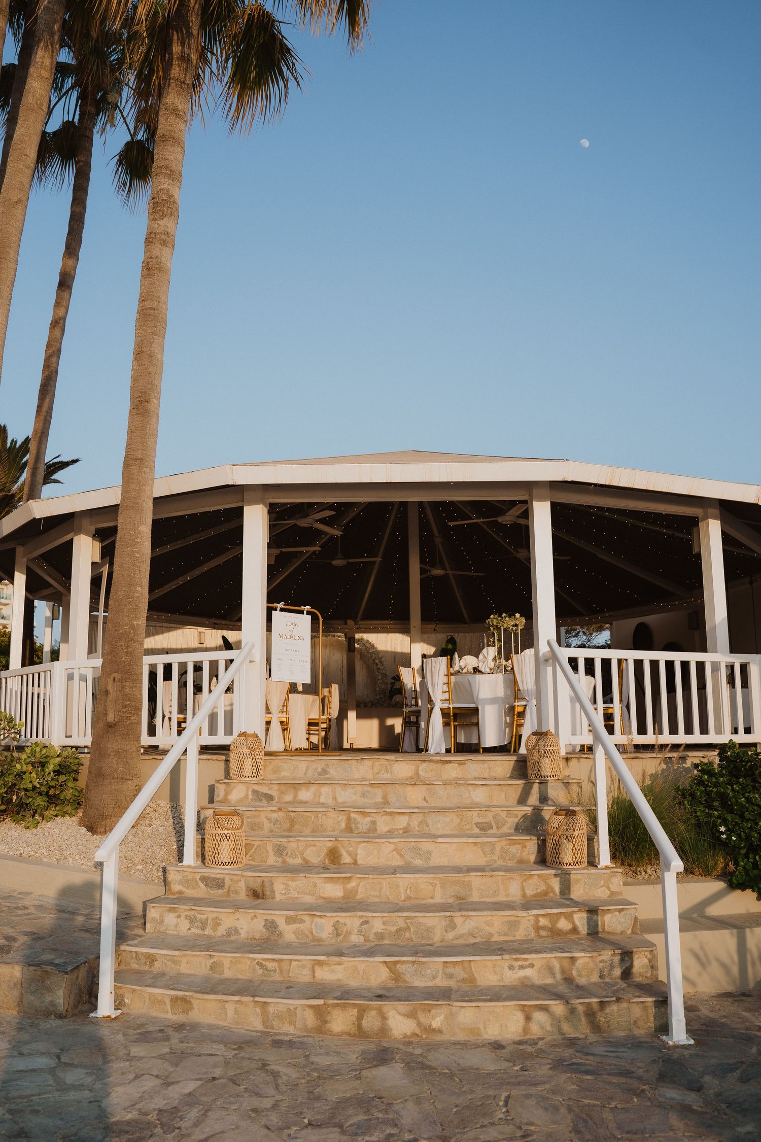 Elegant beachside wedding venue pavilion with white columns, palm trees, and stone steps at sunset.