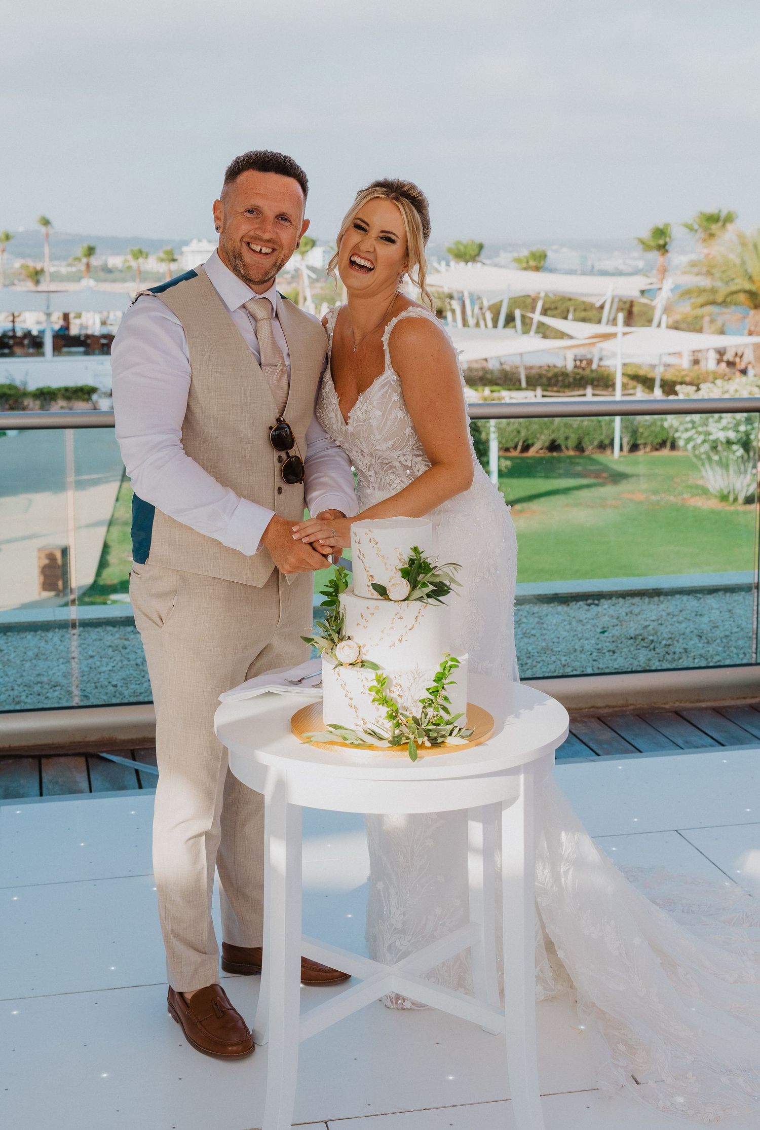 A couple in wedding attire laughs while cutting their cake together at an outdoor waterfront venue.