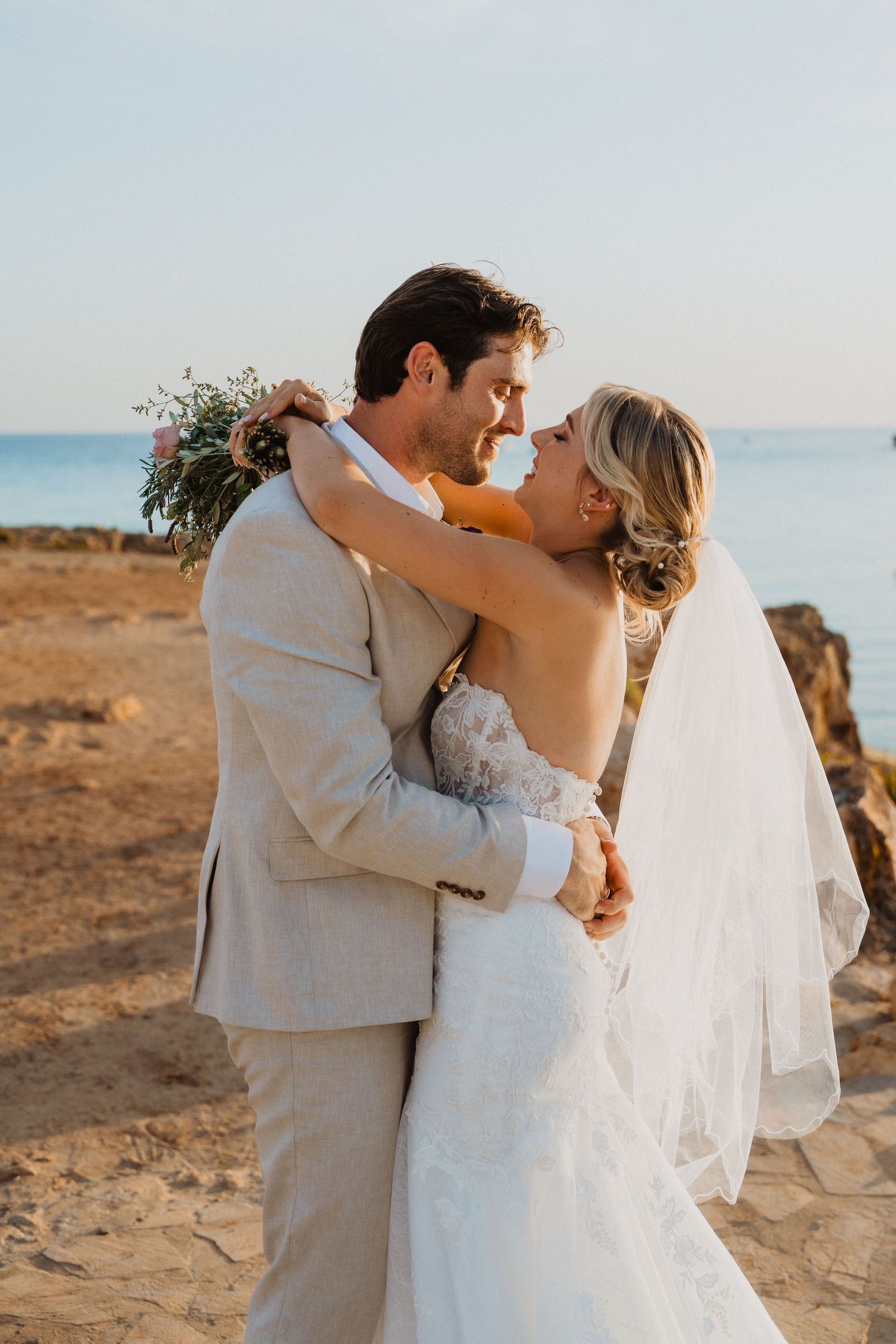 Sunset wedding portrait of couple embracing with ocean view and bouquet.