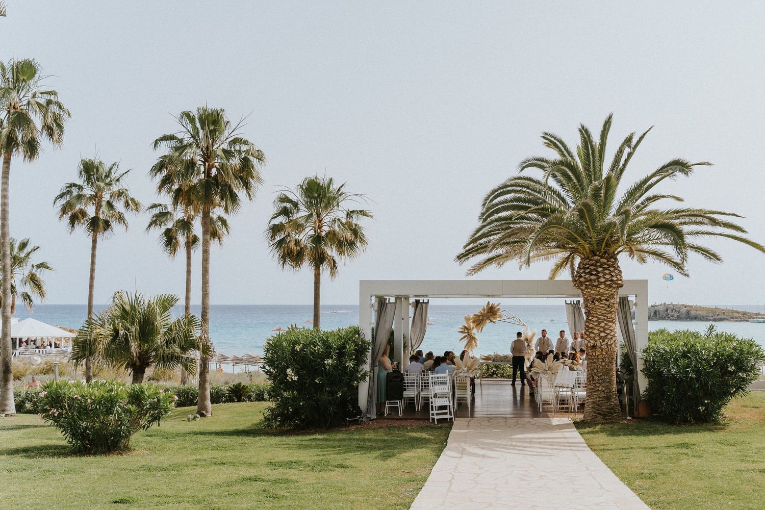 Scenic beachfront wedding ceremony setup with palm trees and a white gazebo against an ocean backdrop.