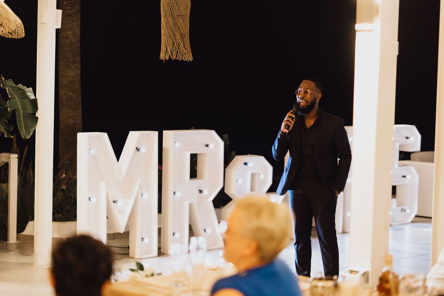 Speaker addresses wedding guests in front of illuminated MR & MRS letters during reception.