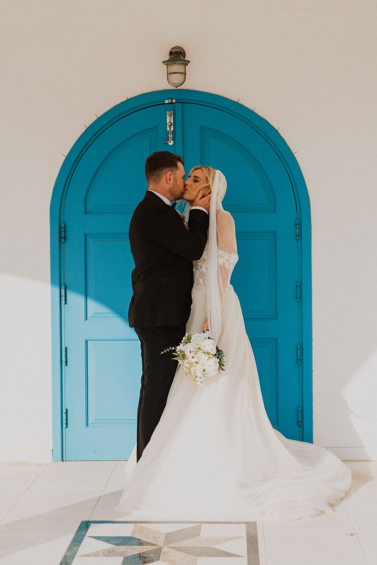 Romantic wedding couple embraces in front of vibrant turquoise church door beneath white arched doorway.