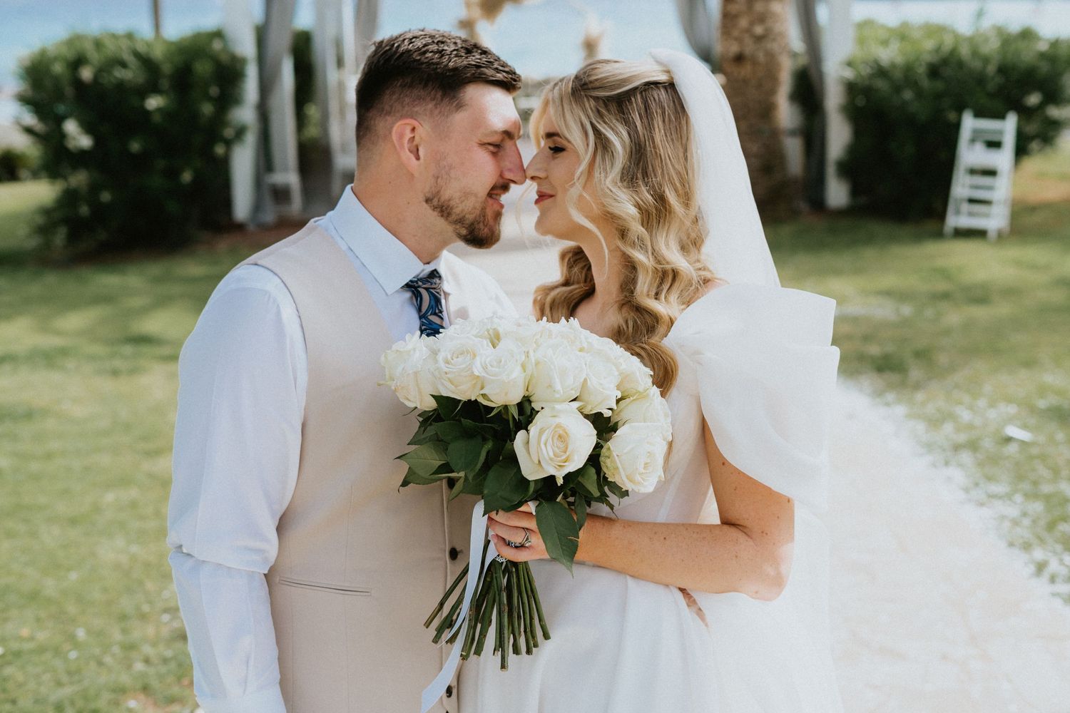 A couple shares an intimate moment at an outdoor wedding ceremony while holding a bouquet of white roses.