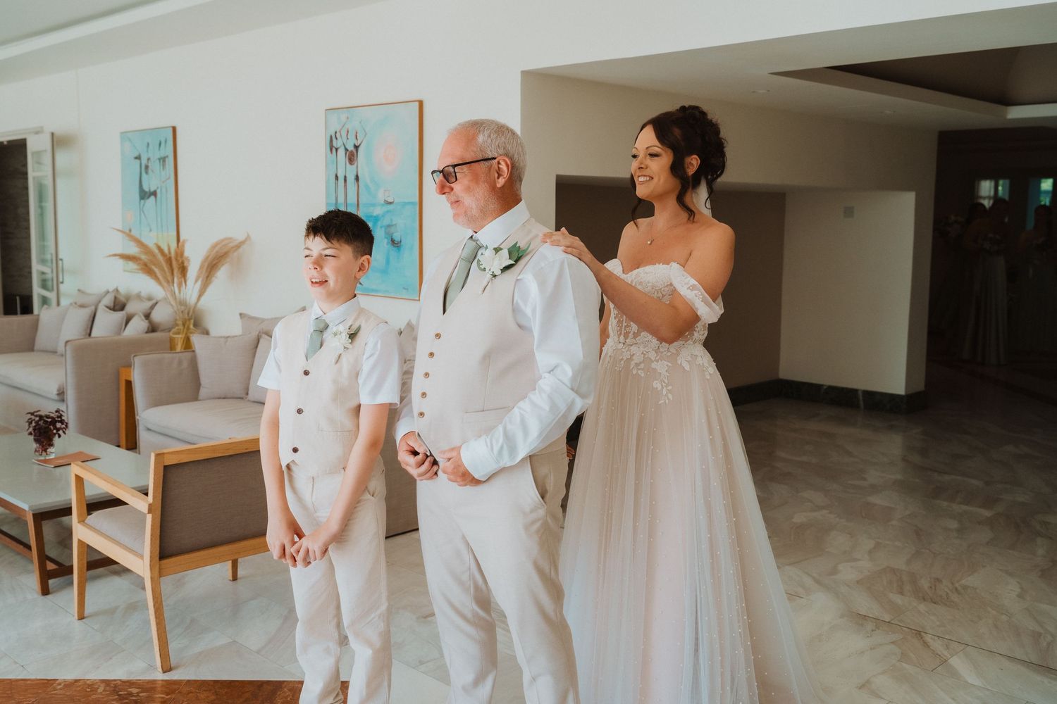 Wedding party members in white attire share a joyful moment before the ceremony in a bright interior space.