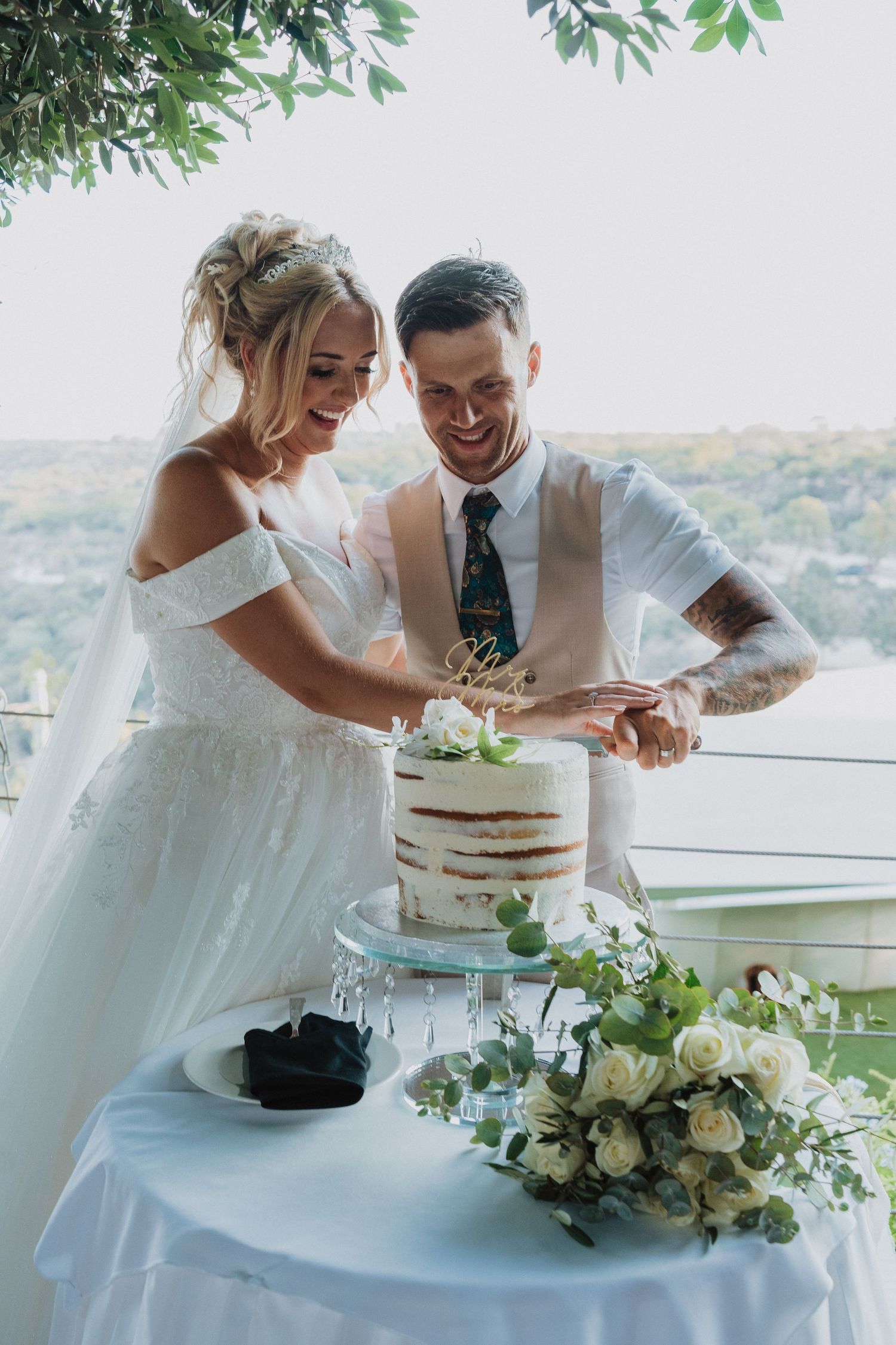 Newlyweds cut their simple white naked cake together at outdoor reception with mountain views.