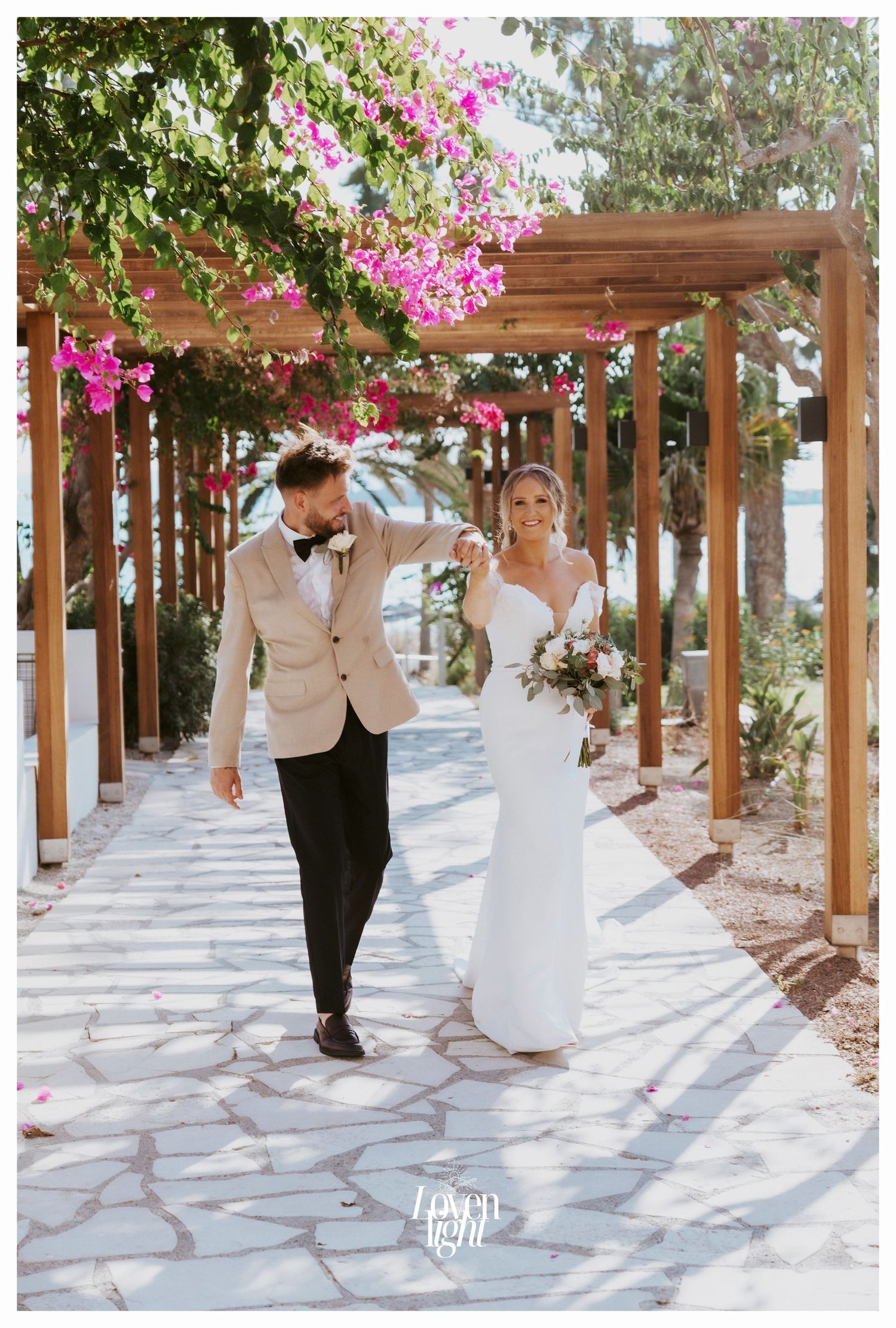 Couple strolls along stone pathway under wooden pergola with cascading pink bougainvillea.