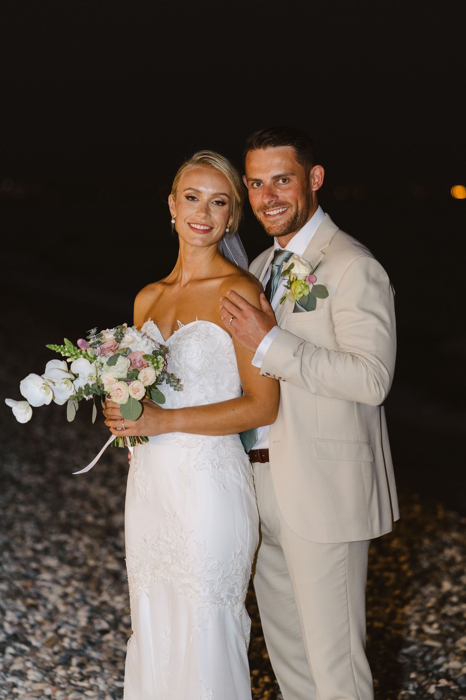 A wedding couple poses together on a beach at night, with bride in white dress holding elegant bouquet.