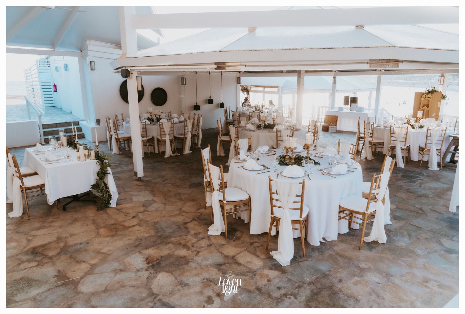 Elegant wedding reception setup with white decor and chiavari chairs in seaside venue.