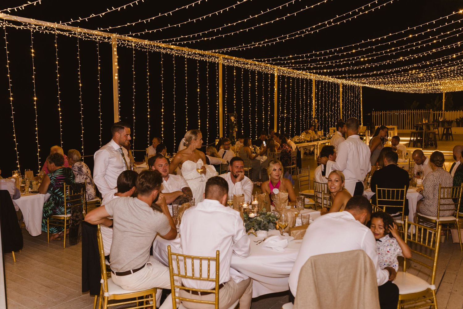 Reception dinner scene with guests dining at elegant tables under string light decor.