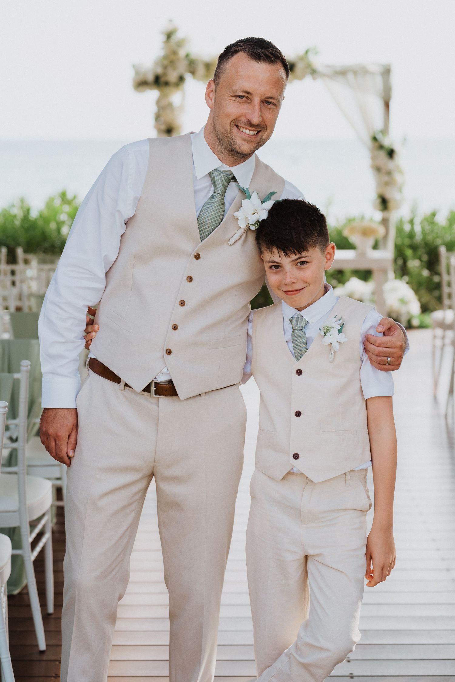A formal outdoor wedding photo showing matching cream-colored suits with sage green ties and boutonnieres.