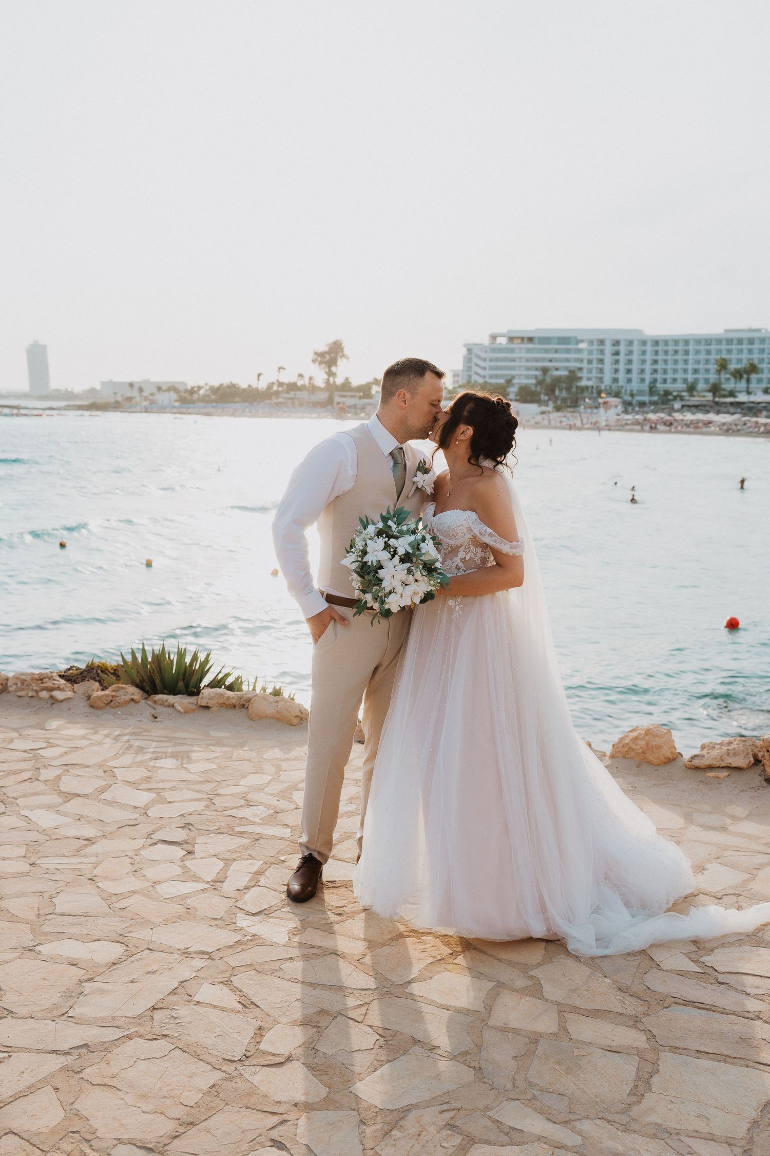 A romantic destination wedding couple share a tender moment on a stone terrace overlooking a Mediterranean beach resort.