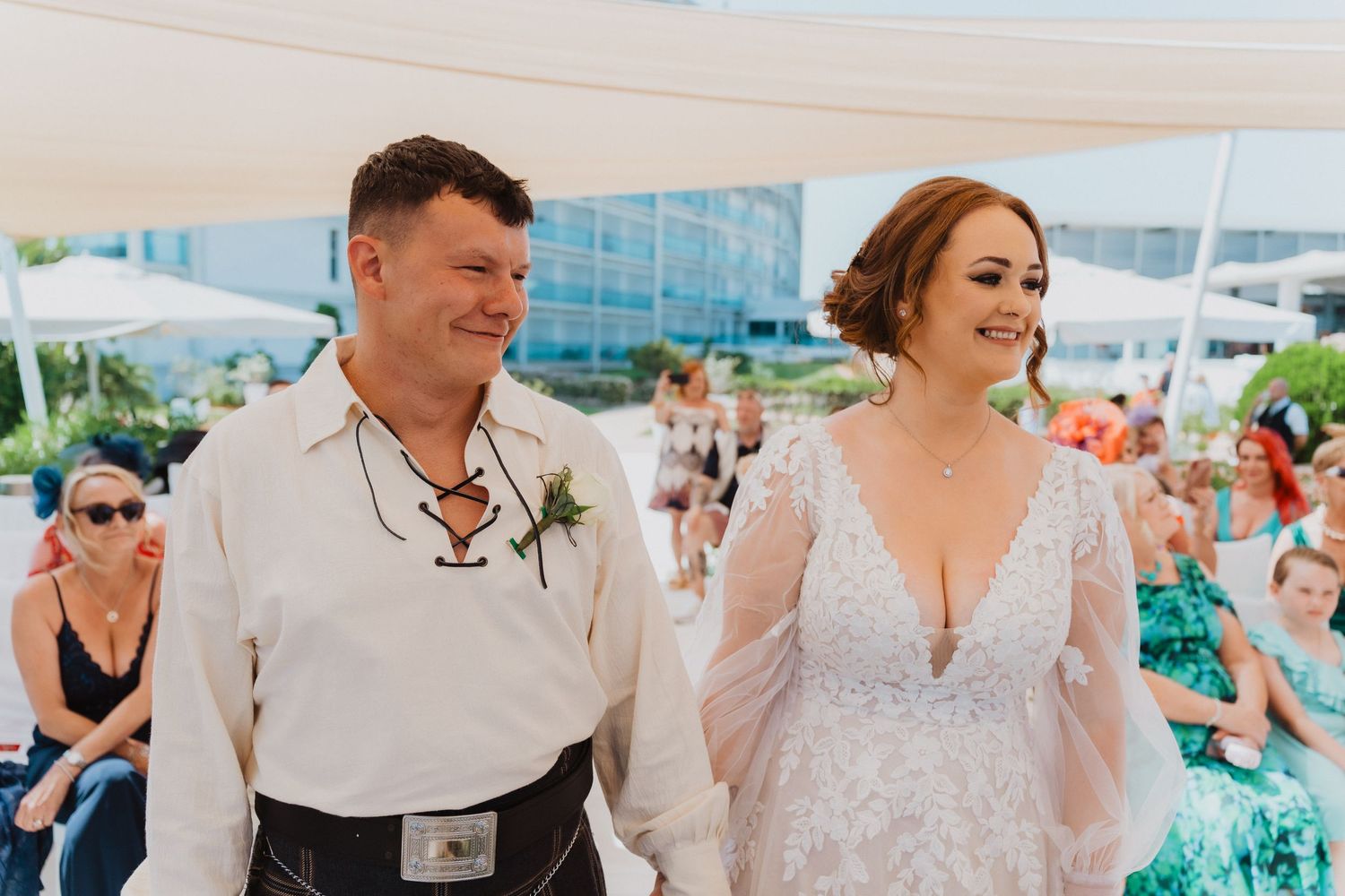 Wedding couple smiles during their beachside ceremony with guests visible in background.