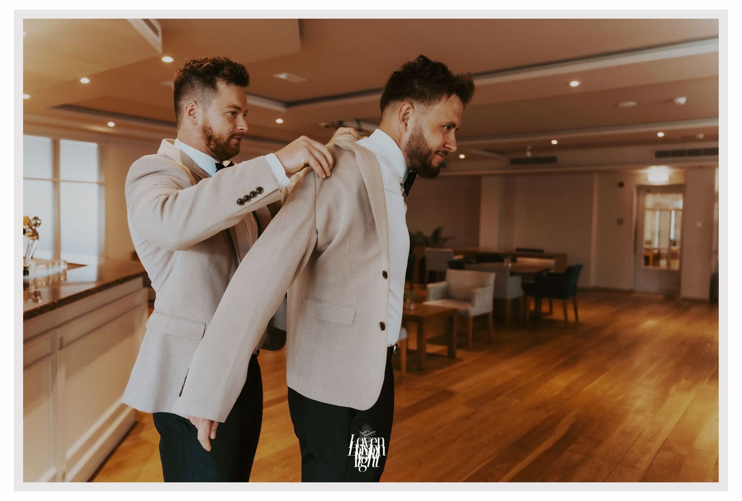 Two groomsmen in beige suits help each other prepare in a wooden-floored room.