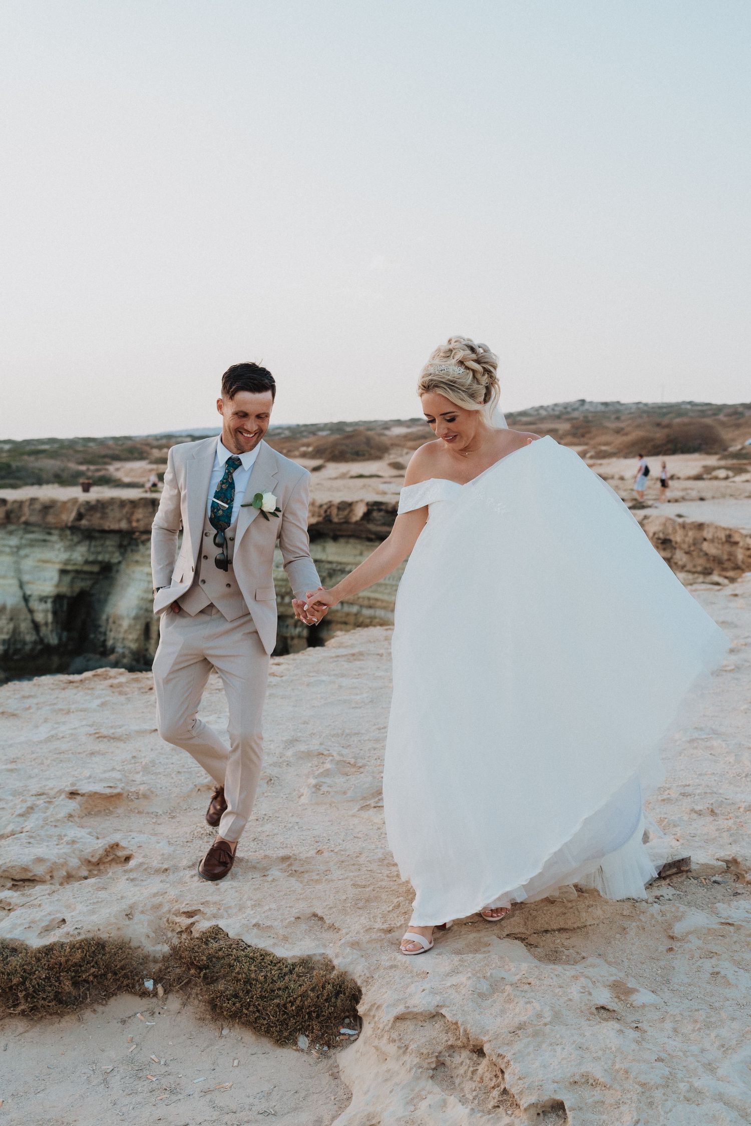 A joyful wedding couple walks together on rocky coastal cliffs overlooking the Mediterranean Sea.