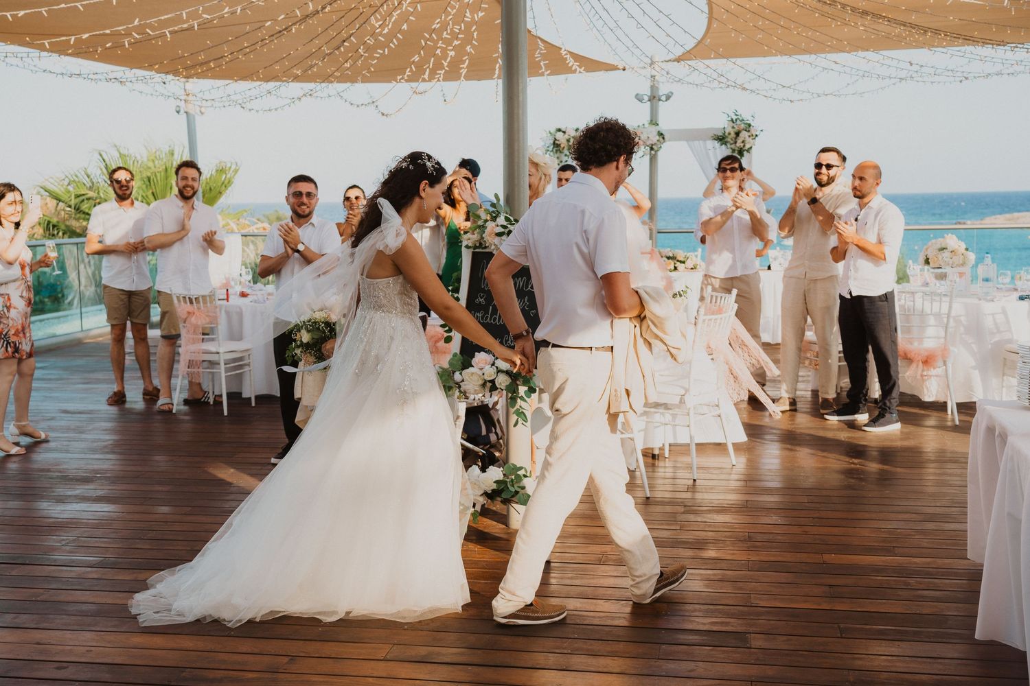 A wedding couple dances together on a wooden deck overlooking the ocean with guests celebrating in the background.