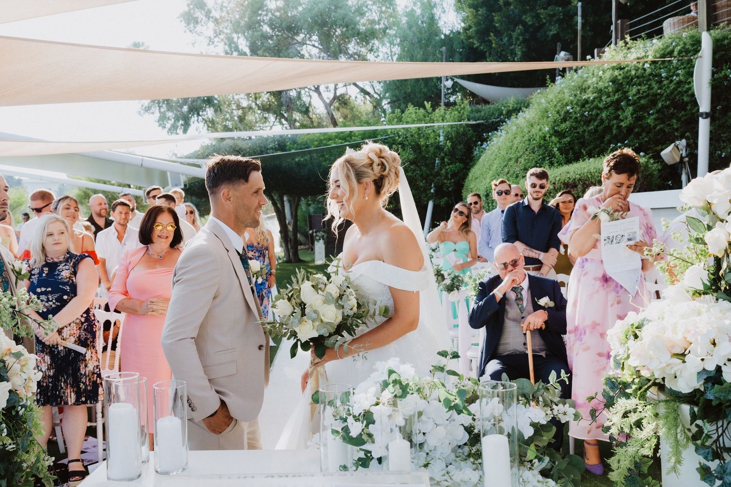 Outdoor wedding ceremony with white floral arrangements and guests watching under shade sails.