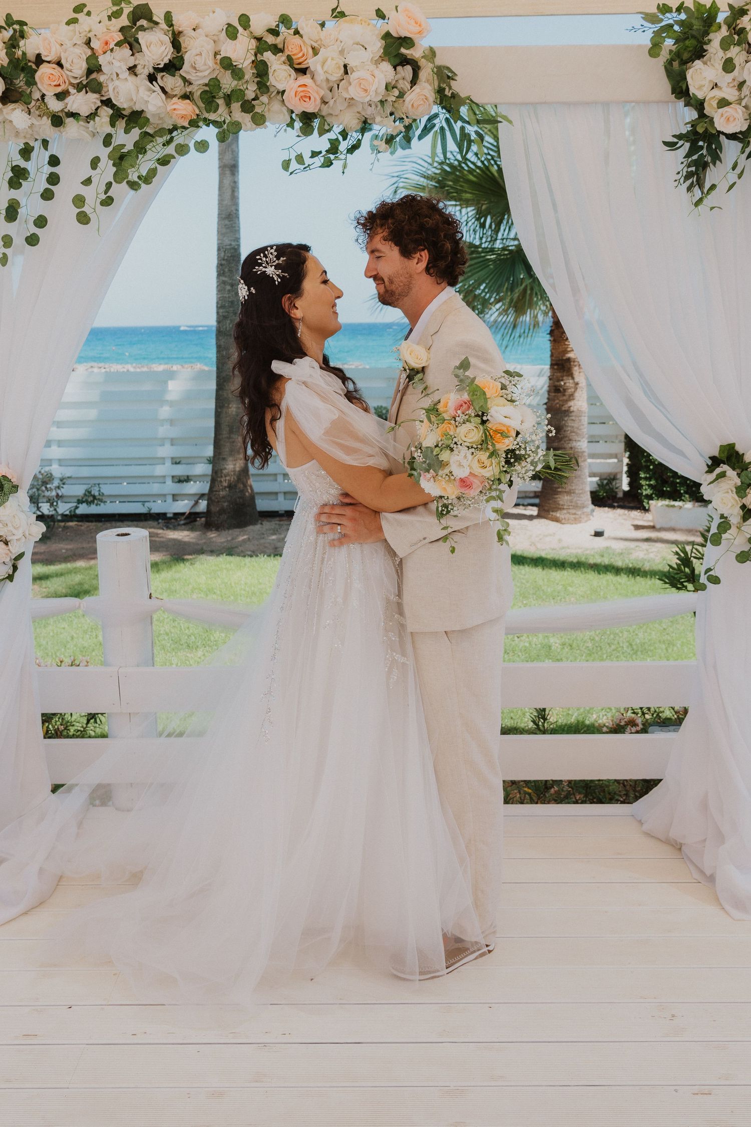 Romantic wedding portrait under floral arch with ocean backdrop and white drapes.