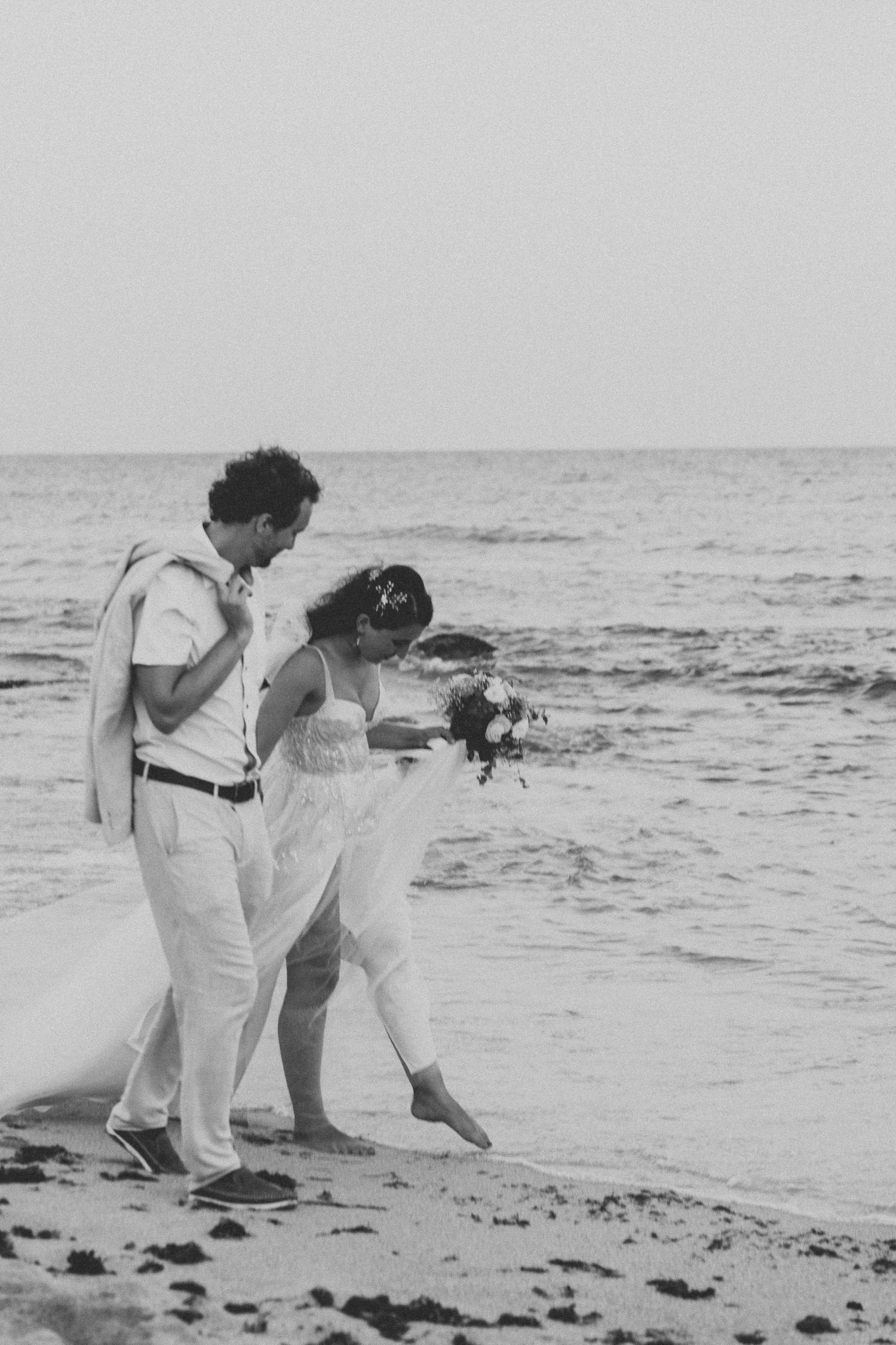 Vintage-style black and white photo of newlyweds walking along beach shoreline.